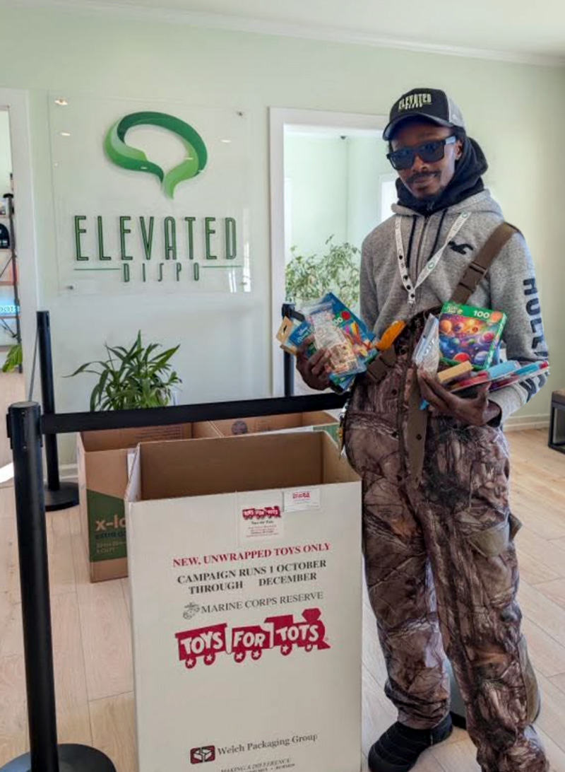 Man holding toys stands next to a Toys for Tots donation box inside Elevated Dispo office.