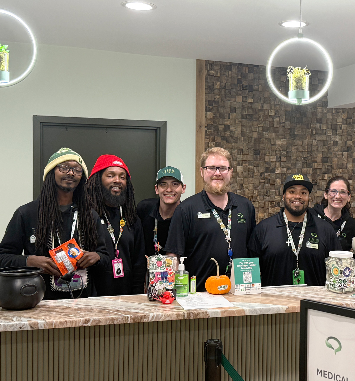 Six smiling employees in branded black shirts stand behind a store counter with various items.