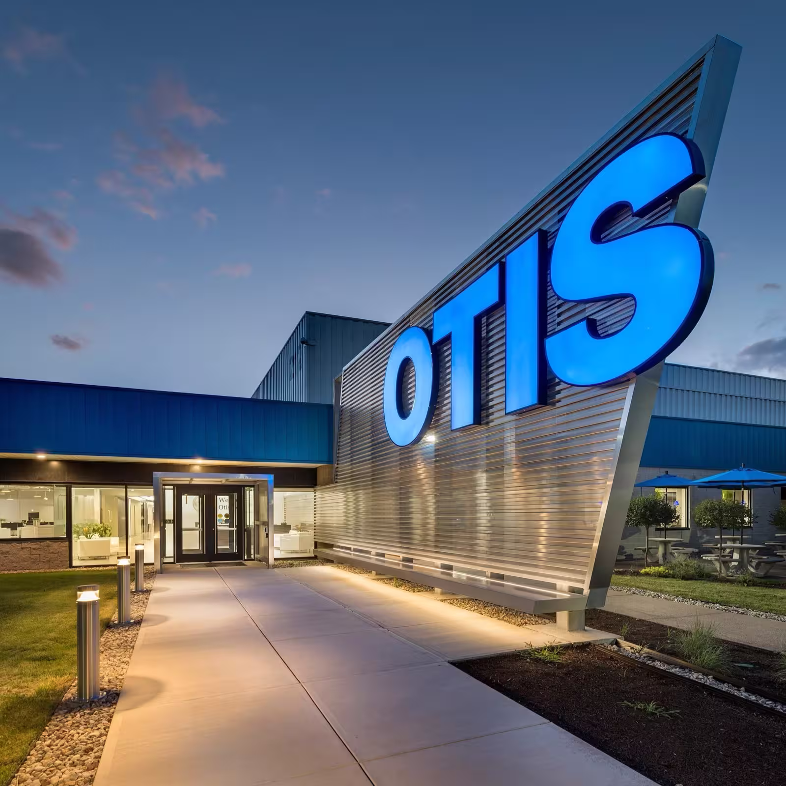 Entrance walkway to a building with large illuminated OTIS sign on metallic wall at dusk.