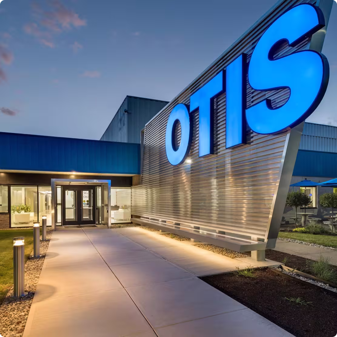 Entrance walkway to a building with large illuminated OTIS sign on metallic wall at dusk.