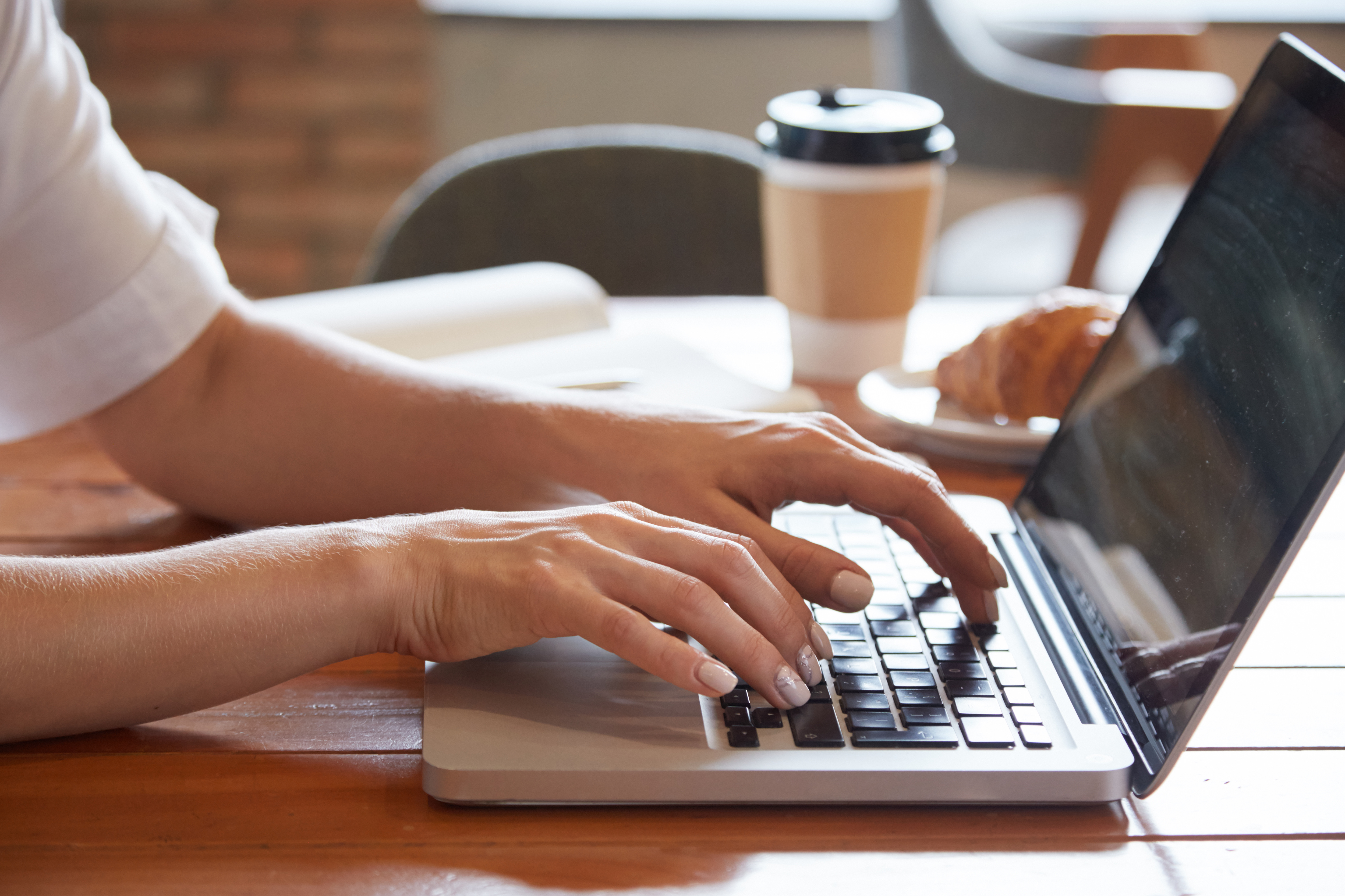 Close-up of hands typing on a laptop keyboard at a wooden table with coffee and croissant nearby.