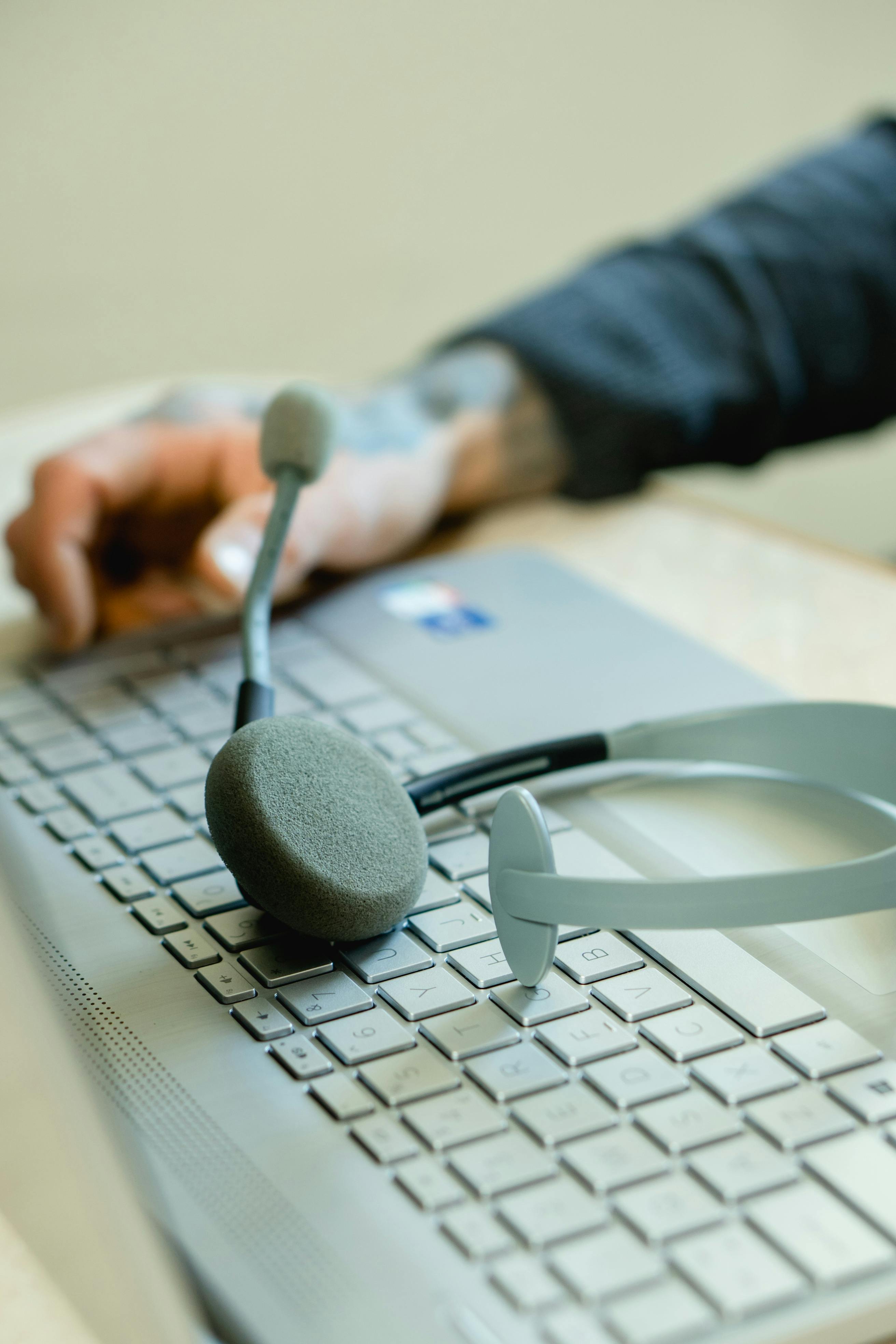 Gray headset with microphone resting on a silver laptop keyboard with a tattooed hand nearby.