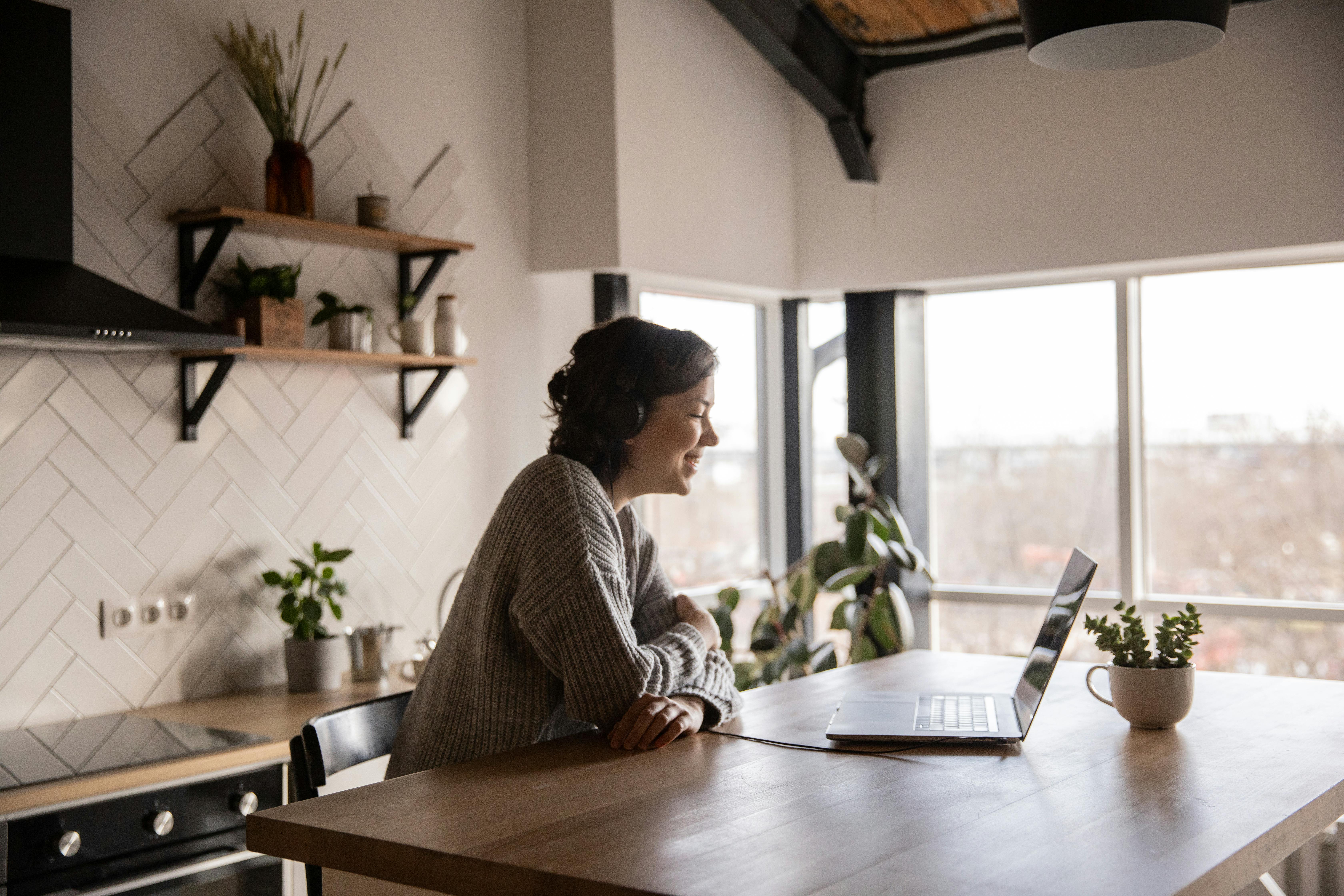 Woman wearing headphones and smiling at laptop while sitting at kitchen table with plants around.