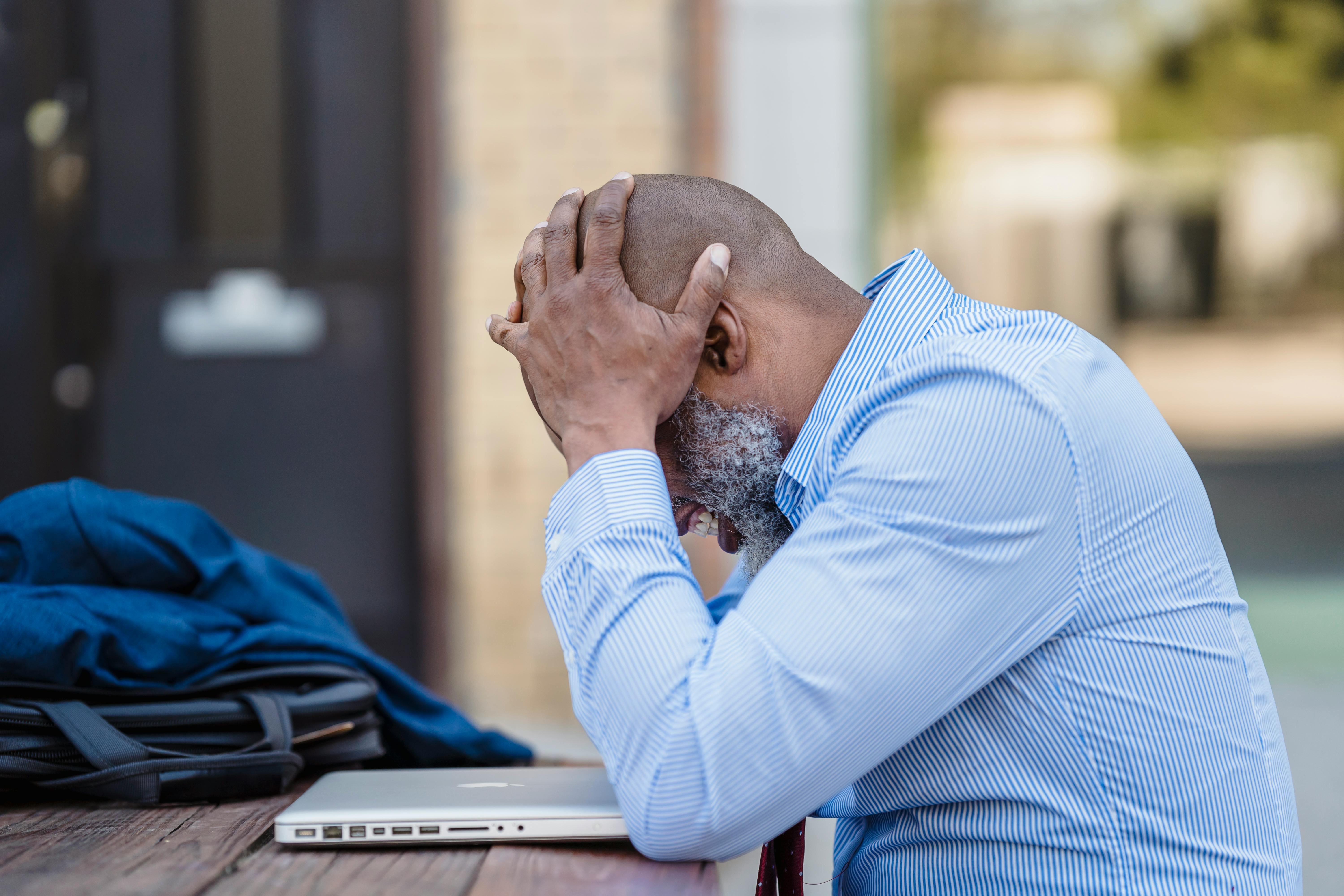 Stressed man holding his head with both hands sitting at a table with closed laptop outdoors.