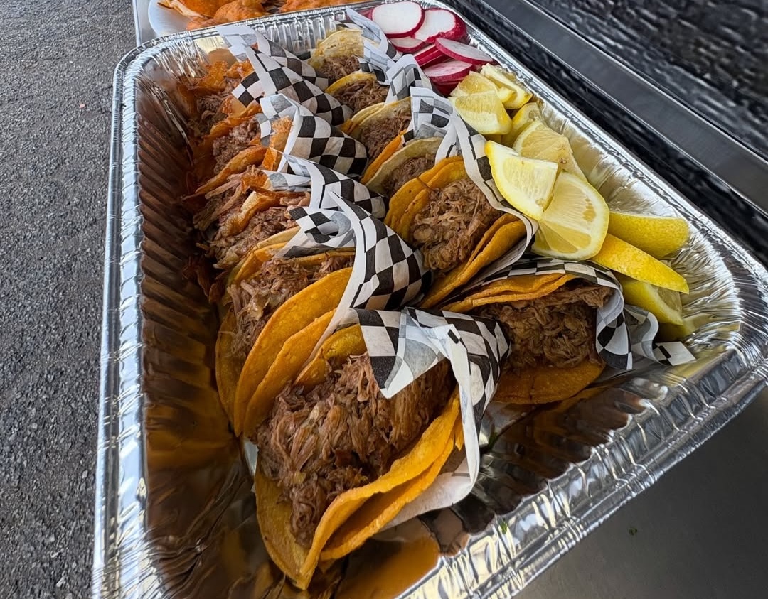 Aluminum tray with several shredded meat tacos wrapped partially in checkered paper, accompanied by sliced radishes and lemon wedges.