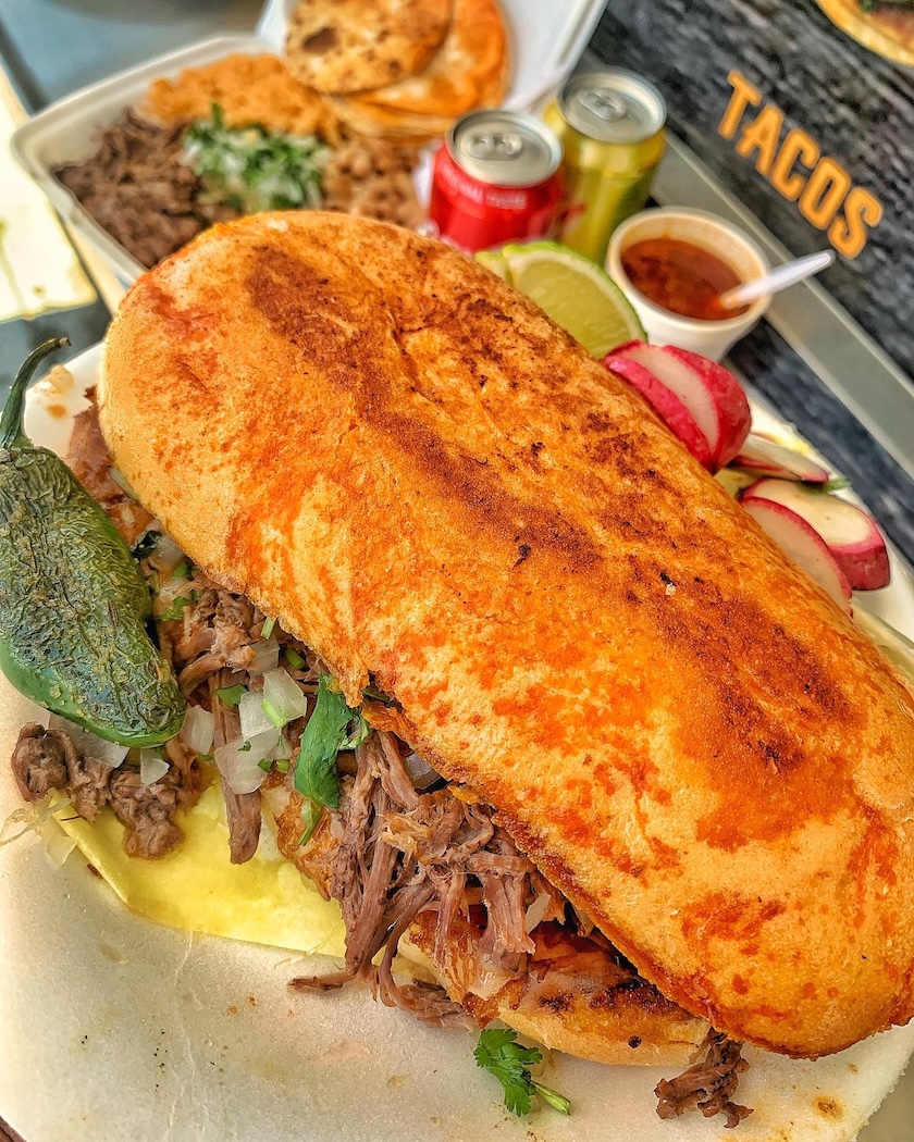 Torta sandwich filled with shredded meat, onions, and cilantro, served with a grilled jalapeño, lime wedge, sliced radishes, salsa, and canned drinks in the background.