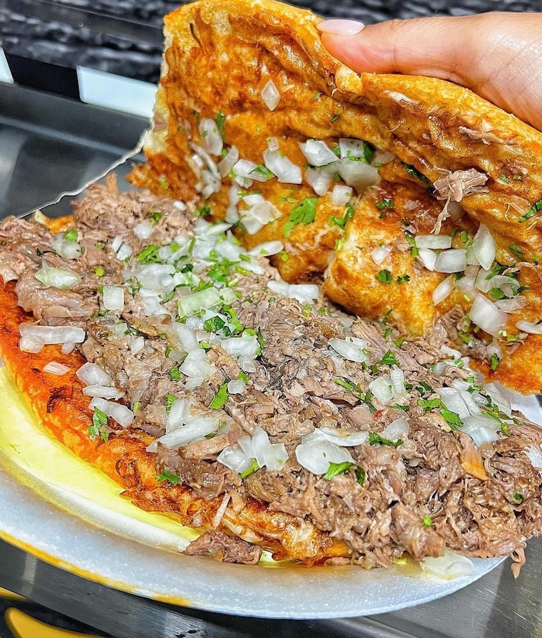Close-up of a hand lifting a crispy torta filled with shredded meat, chopped onions, and cilantro on a white plate.