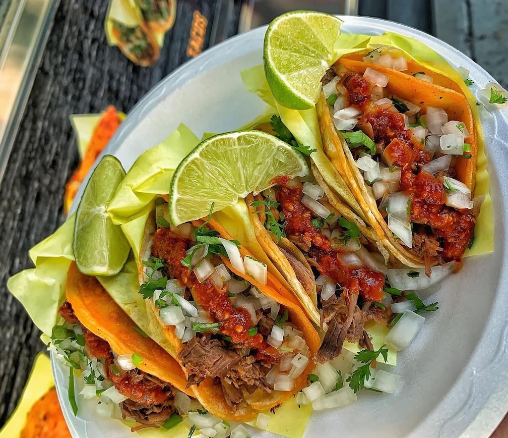 Three tacos with shredded meat, chopped onions, cilantro, red sauce, and lime wedges on a white plate.