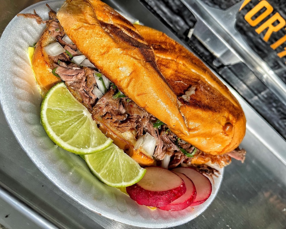Torta sandwich filled with shredded meat, onions, and cilantro on a white paper plate with lime wedges and radish slices.