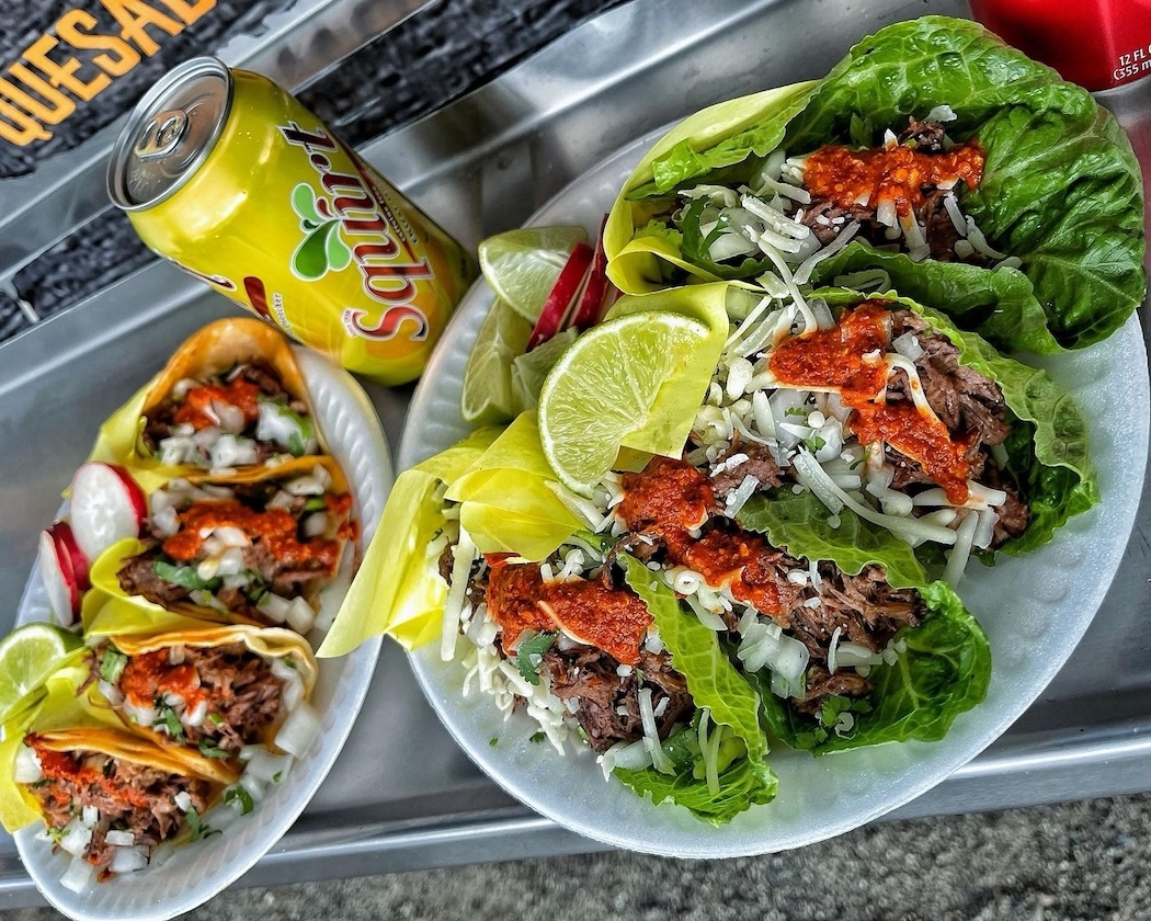 Two plates of tacos: one with three tacos in yellow corn tortillas, and another with lettuce-wrapped tacos topped with cheese and red sauce, alongside lime wedges and a can of Squirt soda.