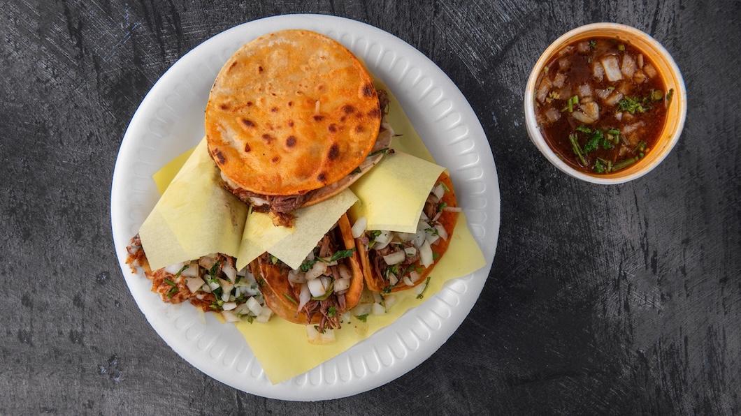 Plate with three tacos topped with onions and cilantro, one taco covered with a toasted tortilla, and a cup of red salsa with chopped onions and cilantro on the side.
