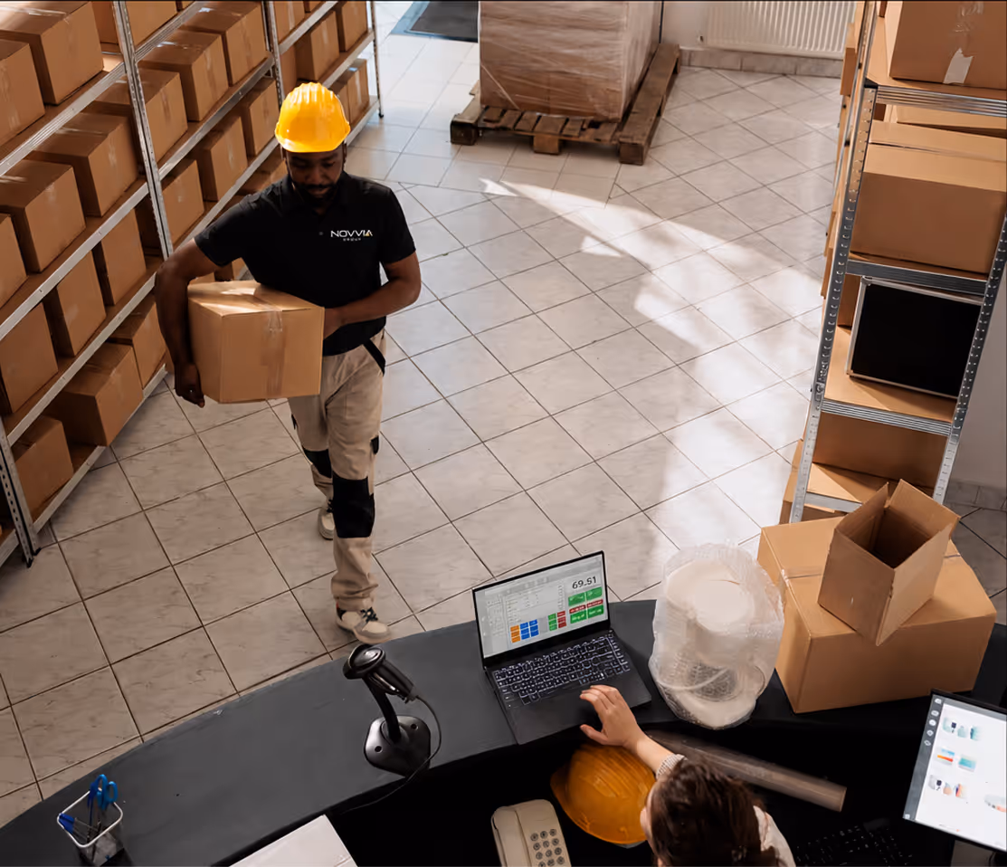 Warehouse worker carrying a box, while another checks laptop at a curved desk with boxes around.