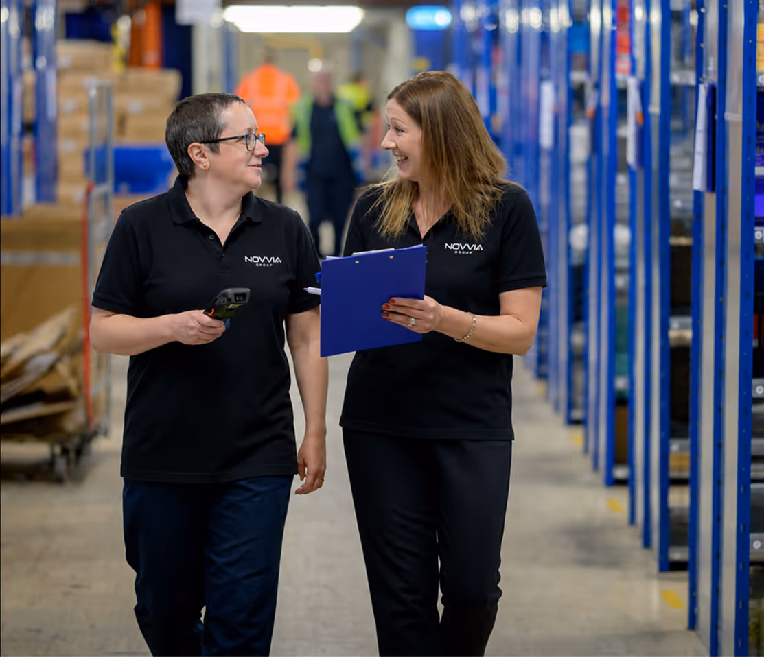 Two women in black Novvia Group shirts talking and walking in a warehouse aisle with shelves and boxes.