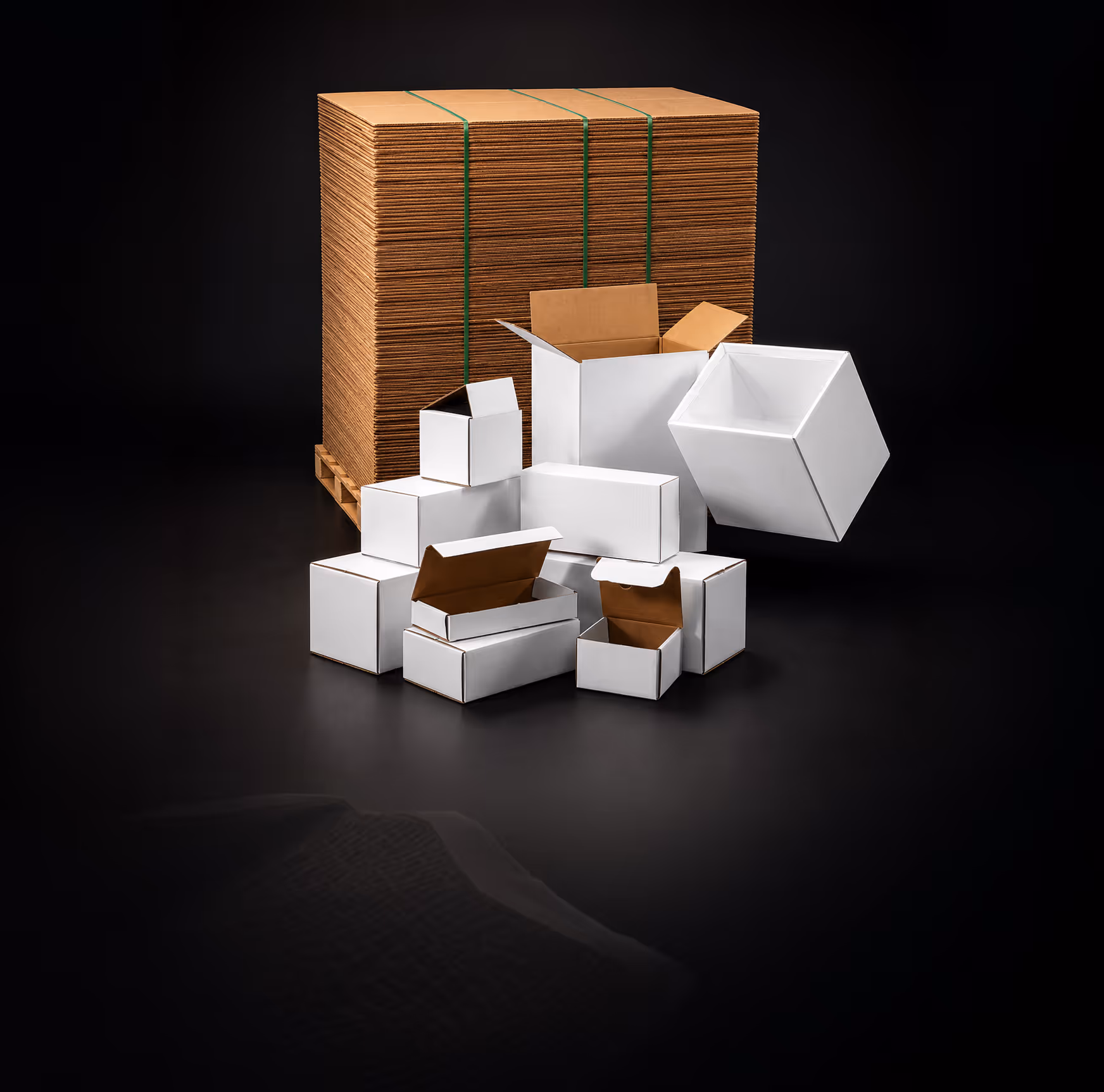 Stack of flattened cardboard boxes on pallet and various white open and closed boxes on black background.