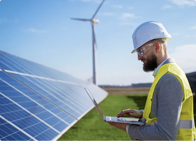 Mann mit Schutzhelm und Warnweste arbeitet an Laptop vor Solarzellen und Windrad im Freien.