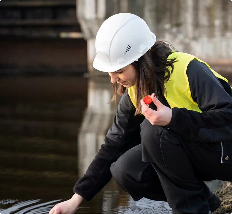 Frau mit Schutzhelm und gelber Sicherheitsweste untersucht Wasserprobe an einem Ufer.