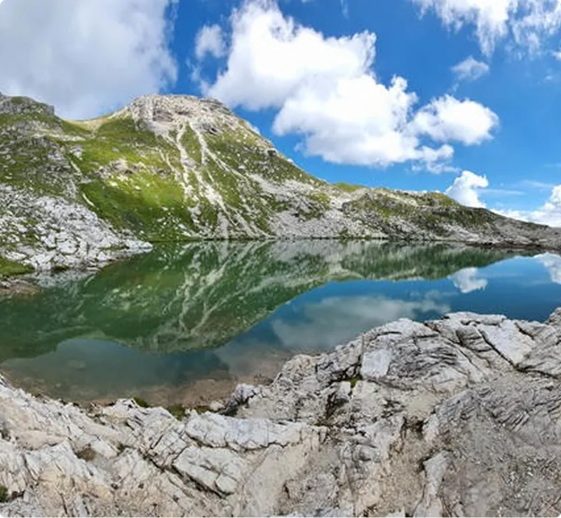 Klarer Bergsee, der die umgebenden felsigen, grünen Hügel und den blauen Himmel mit Wolken spiegelt.