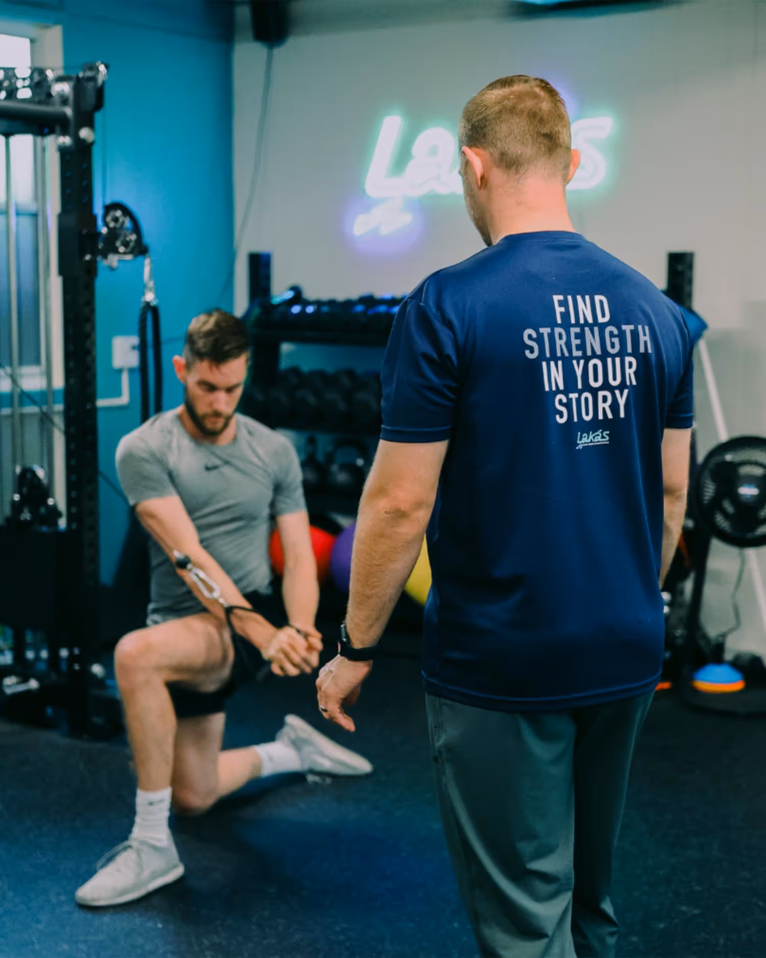 Man in grey shirt exercising with cable machine while another man in navy blue shirt with 'Find Strength in Your Story' text watches in a gym.