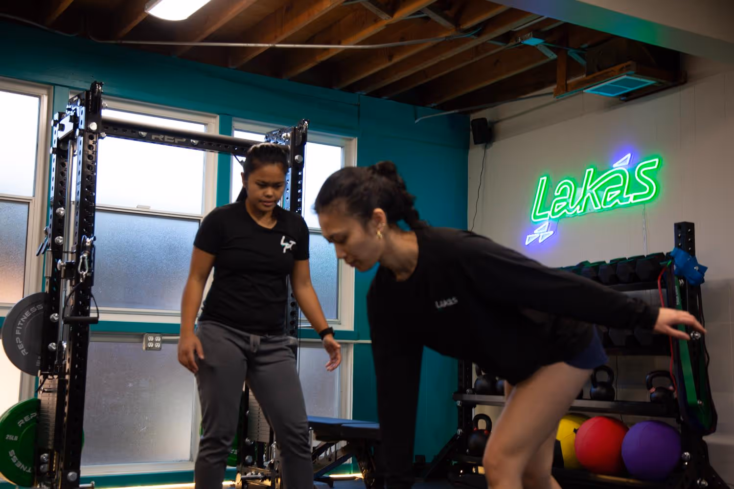 Two women in a gym with one coaching the other performing a bent-over exercise near weights and equipment, with a neon 'Lakas' sign on the wall.