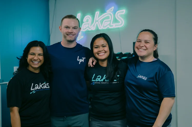 Four smiling people standing close together indoors, wearing Lakás branded shirts with a neon Lakás sign in the background.