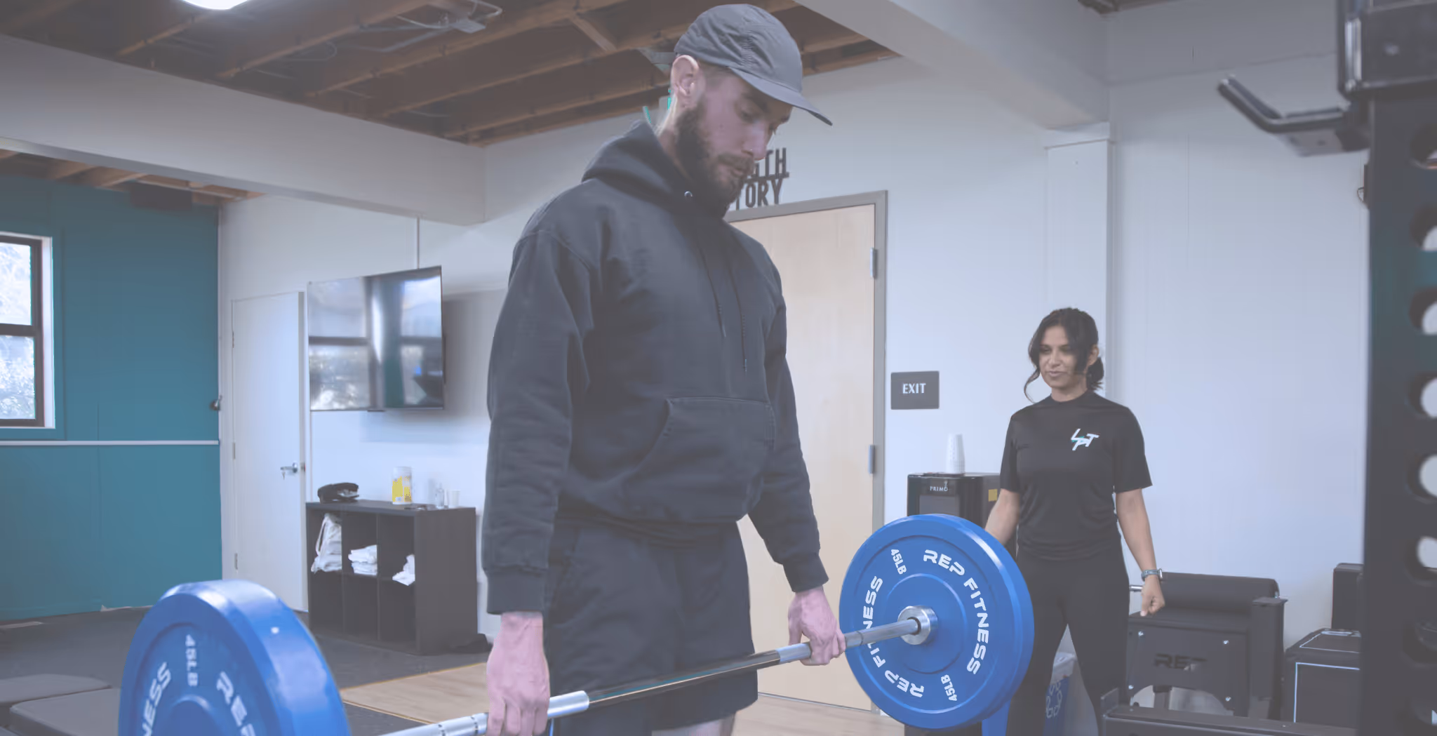 Man in black hoodie lifting a barbell with blue weights in a gym while a woman in black watches and coaches.