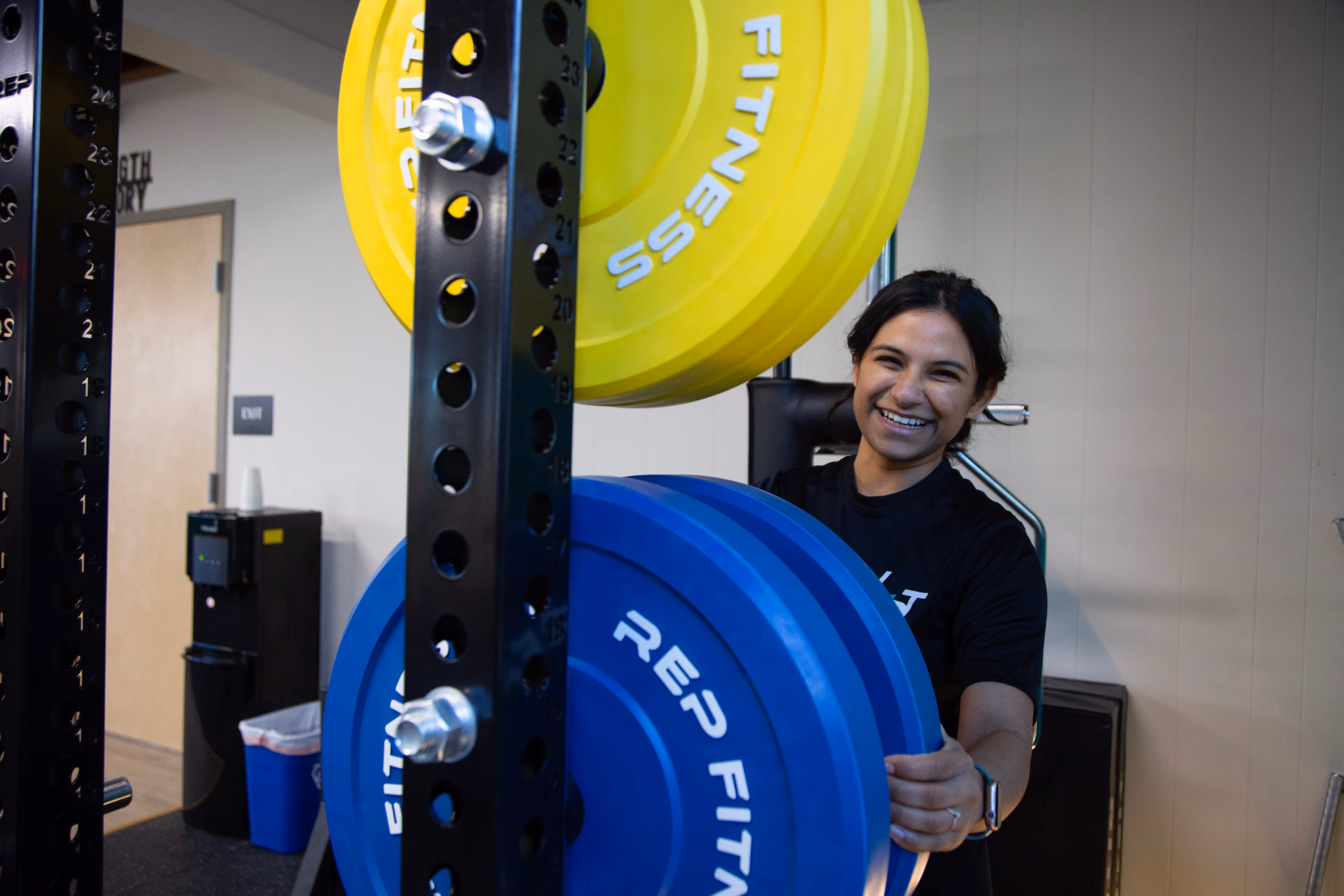 Smiling woman holding large blue and yellow fitness weight plates in a gym.
