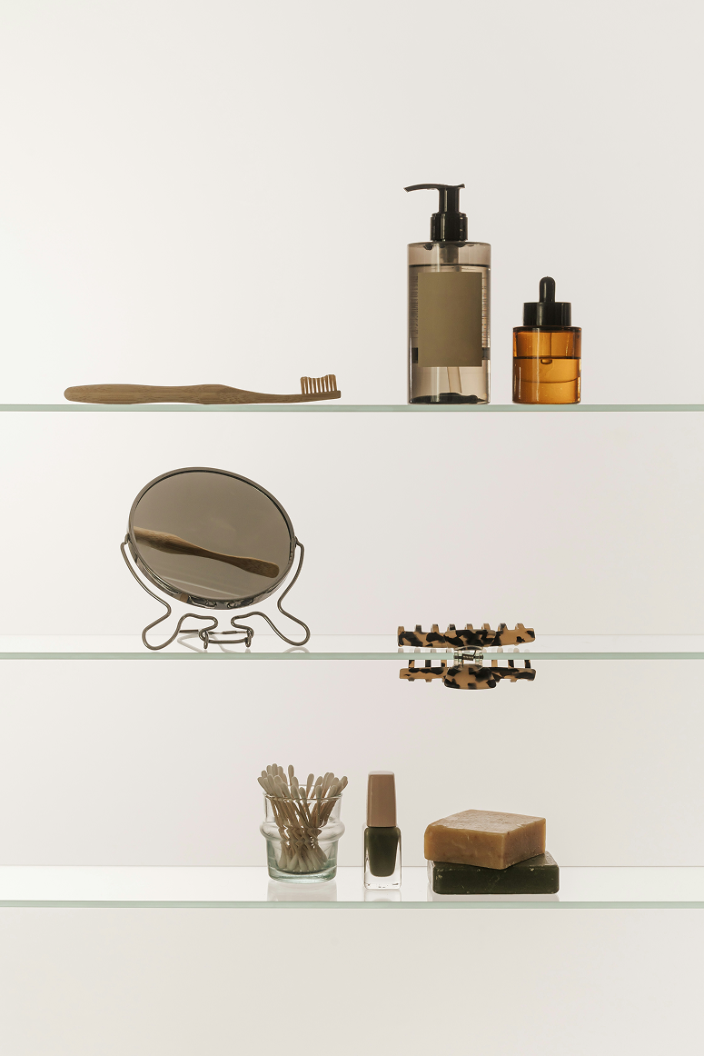 Three glass shelves with personal care items including a wooden toothbrush, pump bottle, dropper bottle, round mirror, hair clip, cotton swabs in a glass jar, nail polish, and two bars of soap.