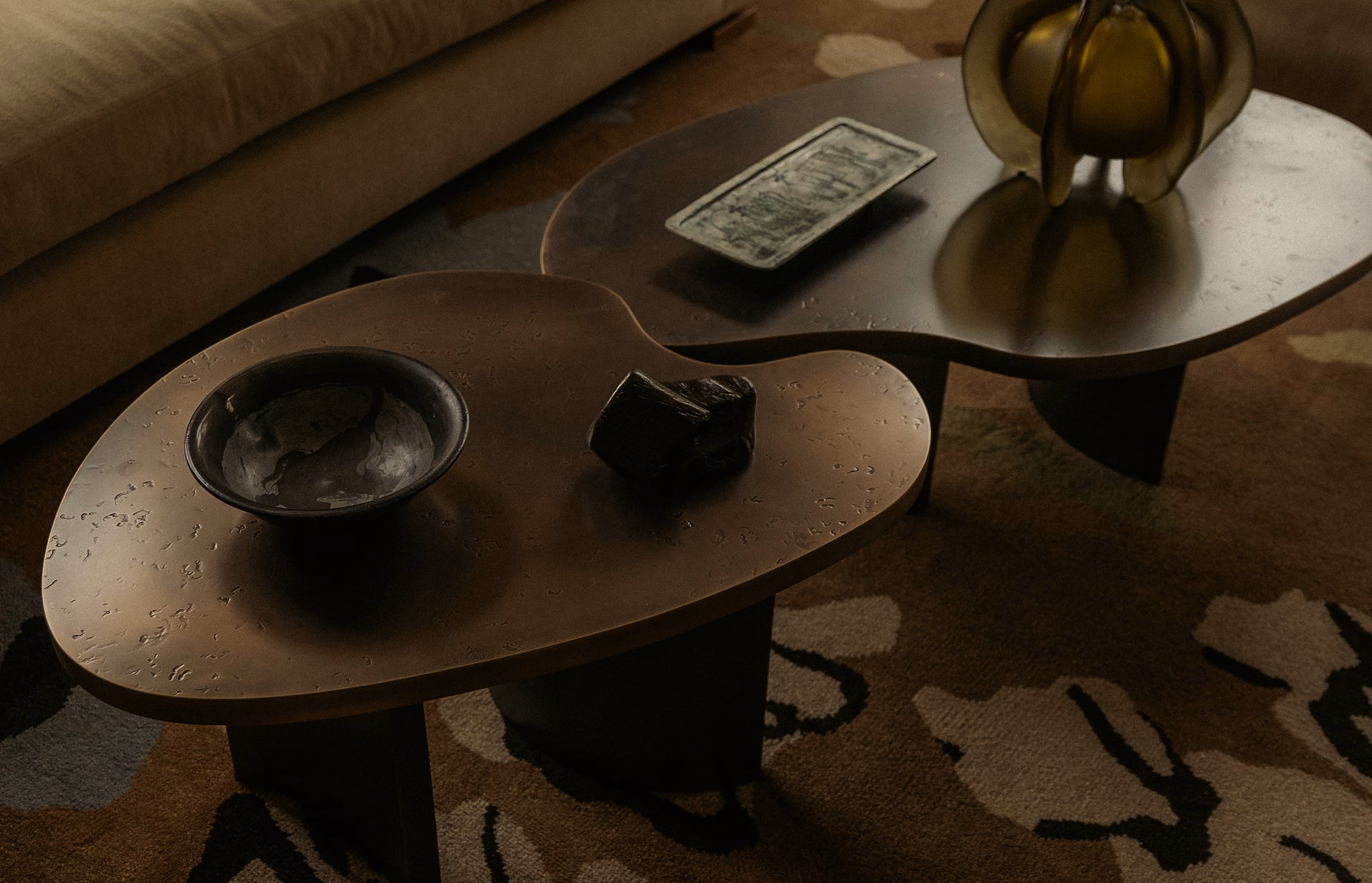 Two irregular wooden coffee tables with dark finish positioned side by side on a patterned carpet; one table holds a small shallow bowl and a dark geometric object, the other has a decorative glass piece and a rectangular blotchy paper item.