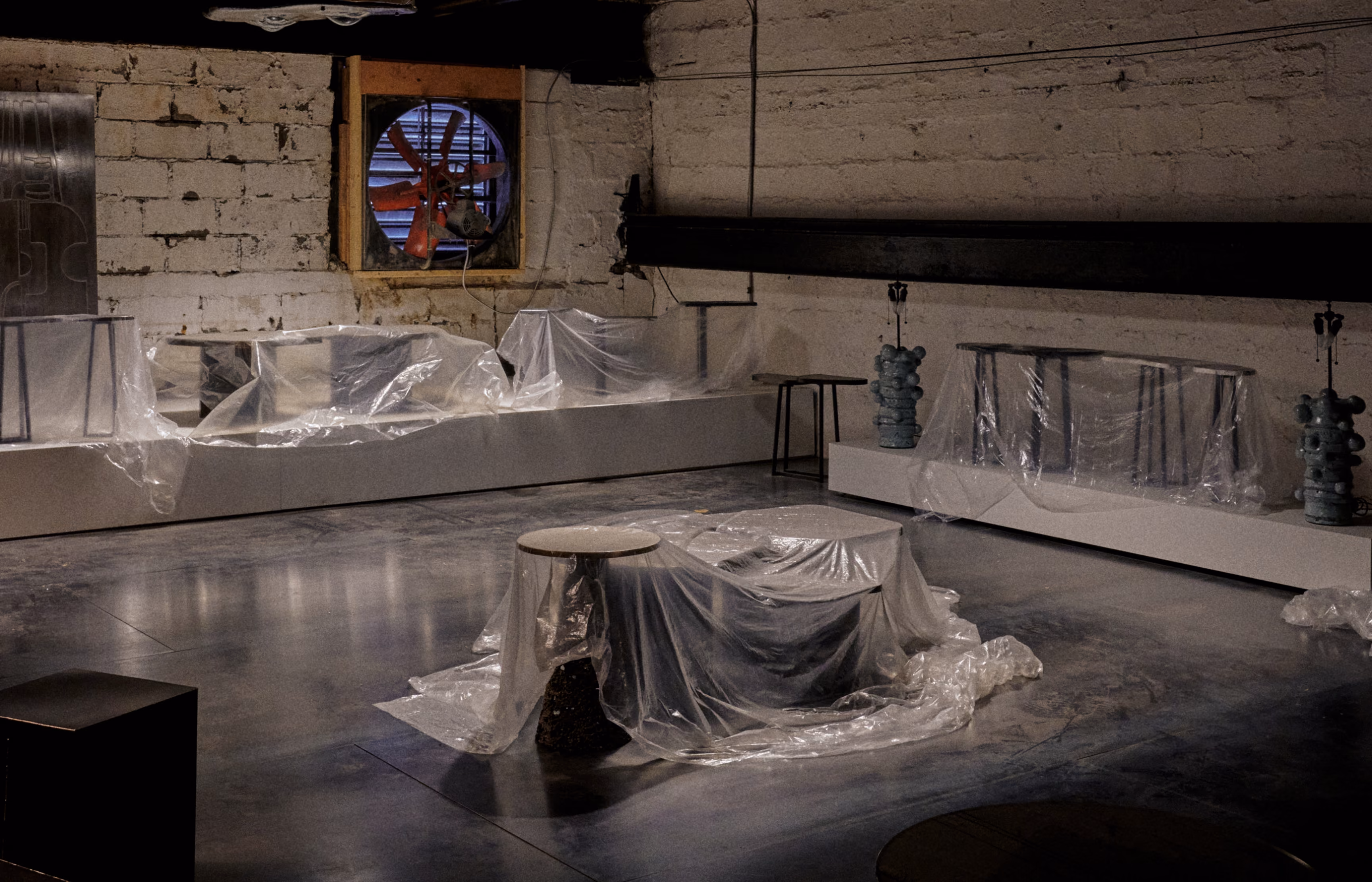 Dimly lit industrial-style room with stone walls and concrete floor, furniture and objects covered in clear plastic sheets, and a small window with a red fan visible outside.