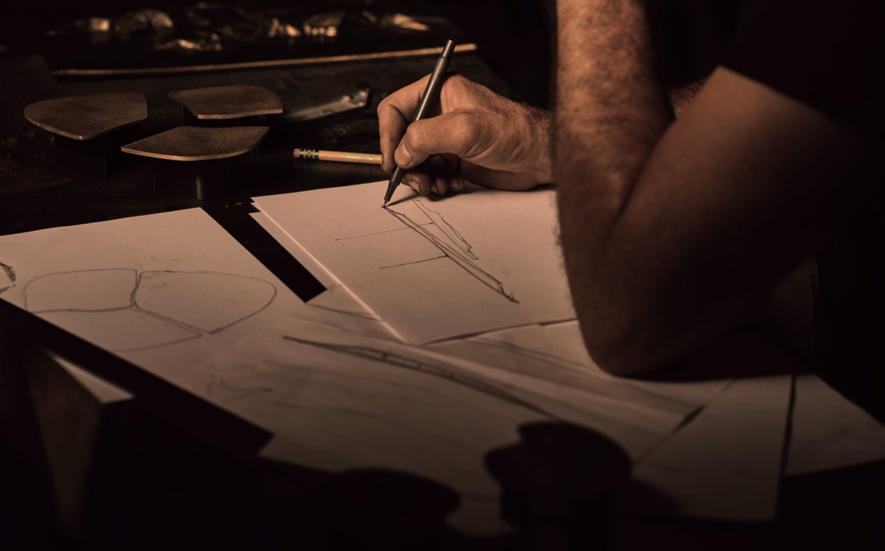 Person with arm resting on table is sketching a design on paper with a pen, surrounded by other sketch papers and pencil in dim light.