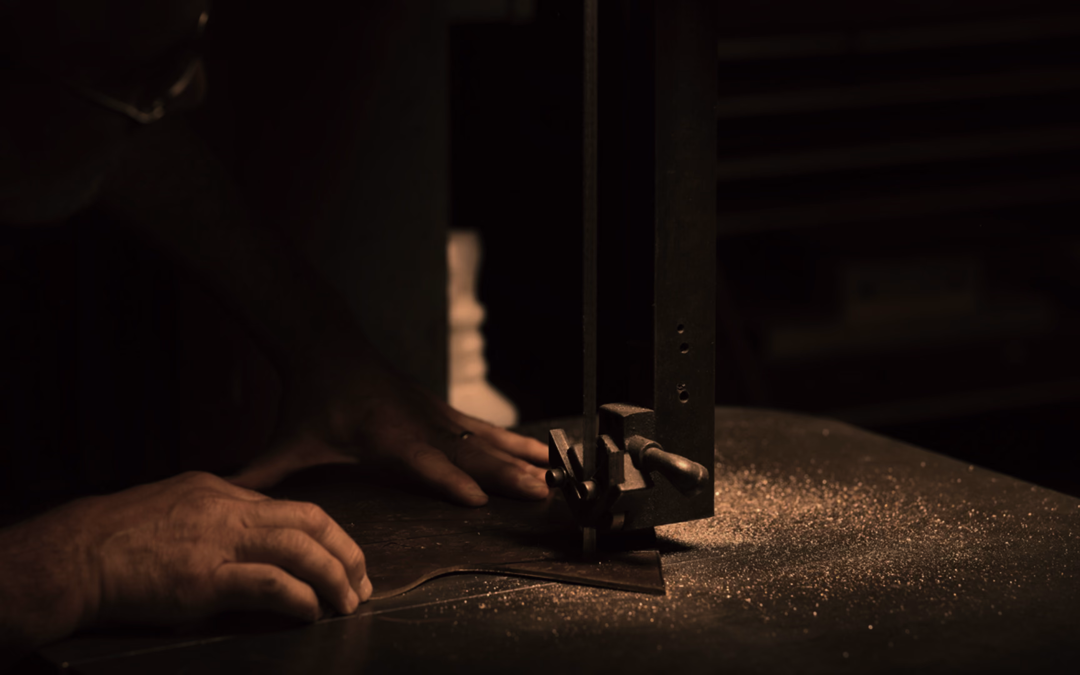Close-up of hands guiding a piece of wood through a bandsaw, producing fine sawdust on a dark work surface in dim light.