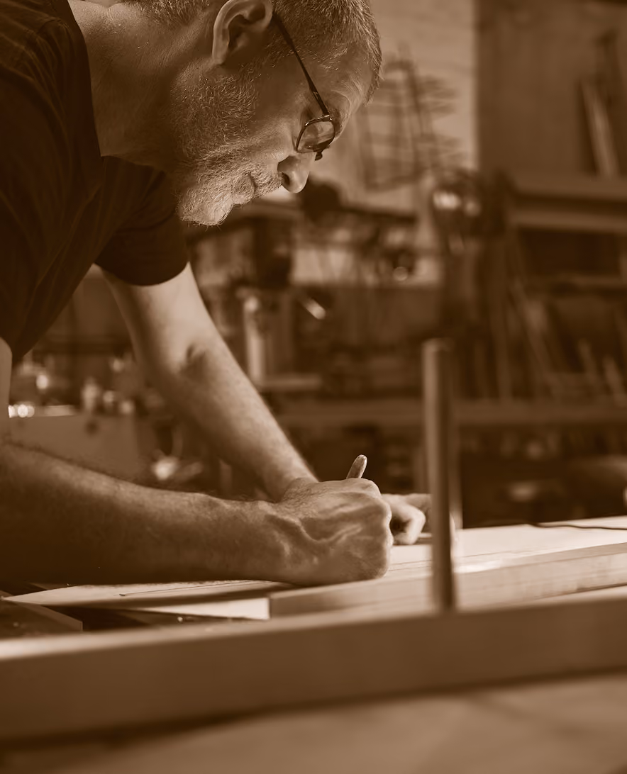 Older man with glasses and a beard leaning over a workbench, drawing or writing on a large sheet of paper in a workshop filled with tools and materials.