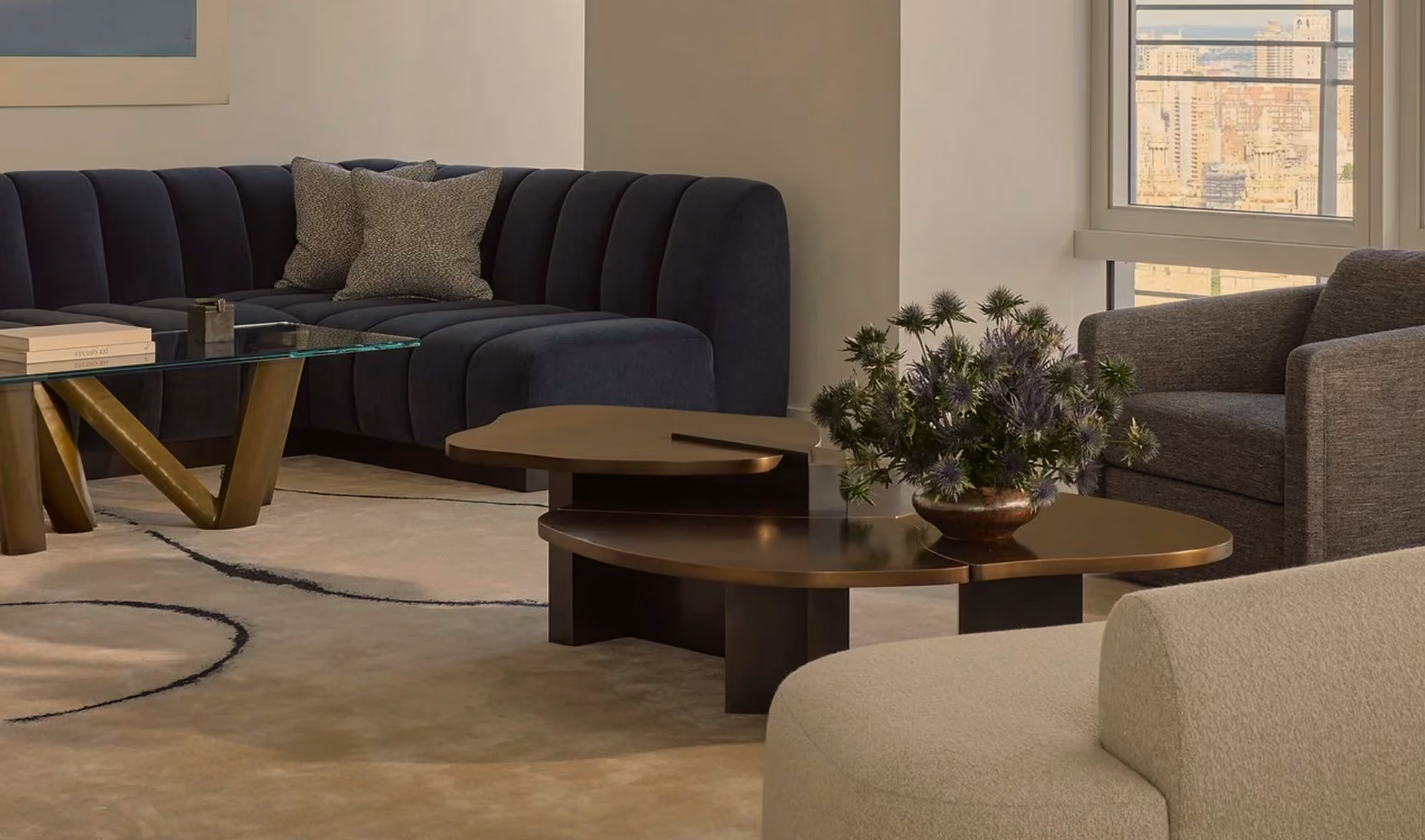 Modern living room featuring a dark blue sectional sofa with cushions, a glass coffee table with gold legs, two nested dark brown round tables with a floral arrangement, a gray armchair, and a beige upholstered chair, with a window showing a cityscape view.