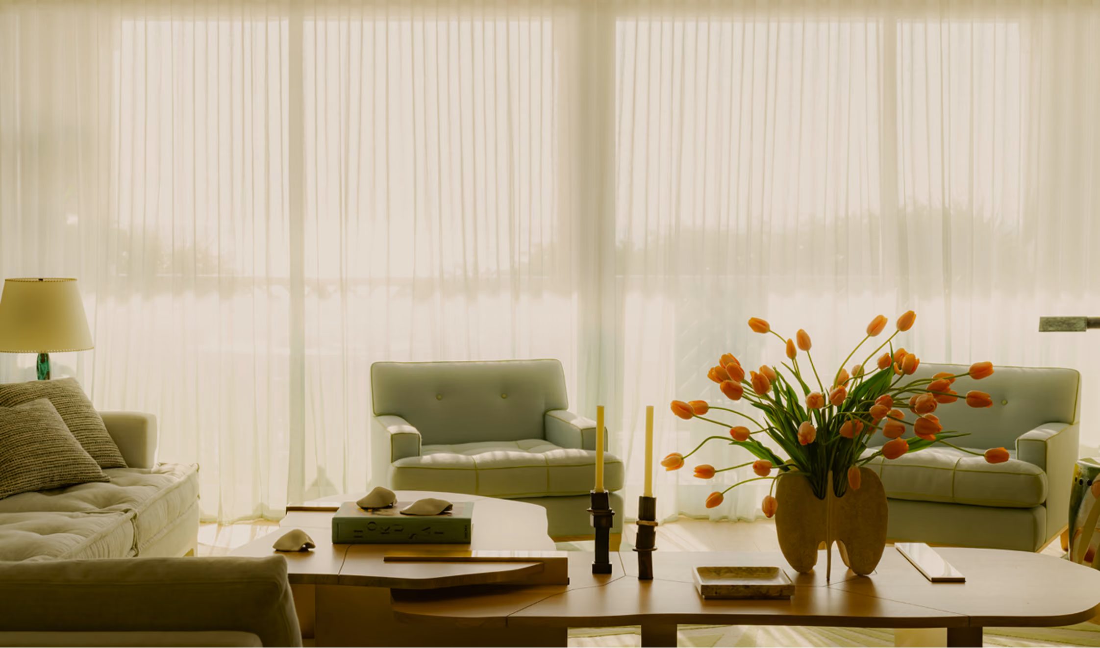 Bright living room with sheer curtains covering large windows, featuring light gray sofas, a wooden coffee table with orange tulips in a vase, candles, books, and a lamp on the side.