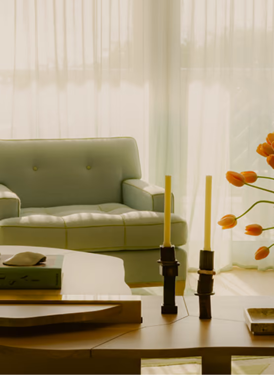 Living room with a light green tufted sofa against sheer white curtains, a wooden coffee table with two tall candles in dark holders, a book, and a vase of orange tulips.