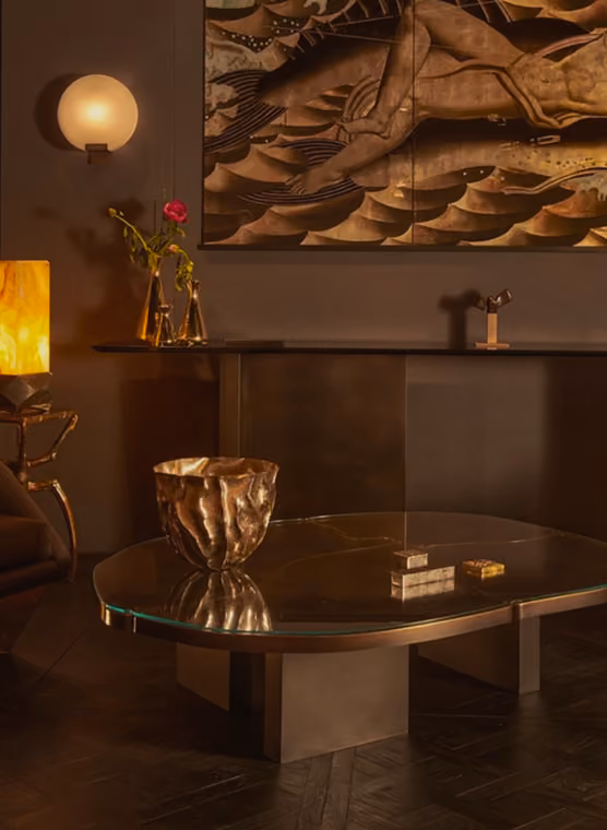 Dimly lit modern living room corner with a glass coffee table featuring a metallic bowl, a console table against the wall with decorative vases holding a single red rose, a sculptural object, a round wall light fixture, and a large artwork depicting abstract human figures and waves.