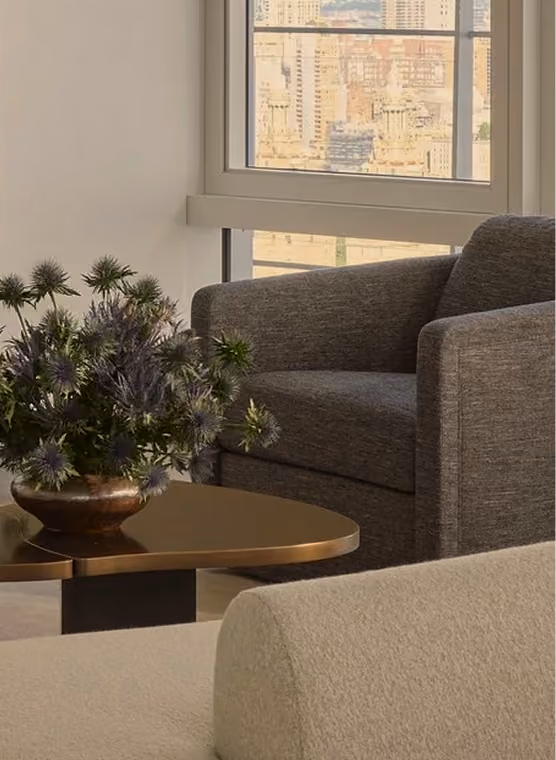 Interior corner showing a modern gray upholstered armchair next to a bronze-colored coffee table with a decorative vase of spiky green and purple flowers, with a window revealing a cityscape in the background.