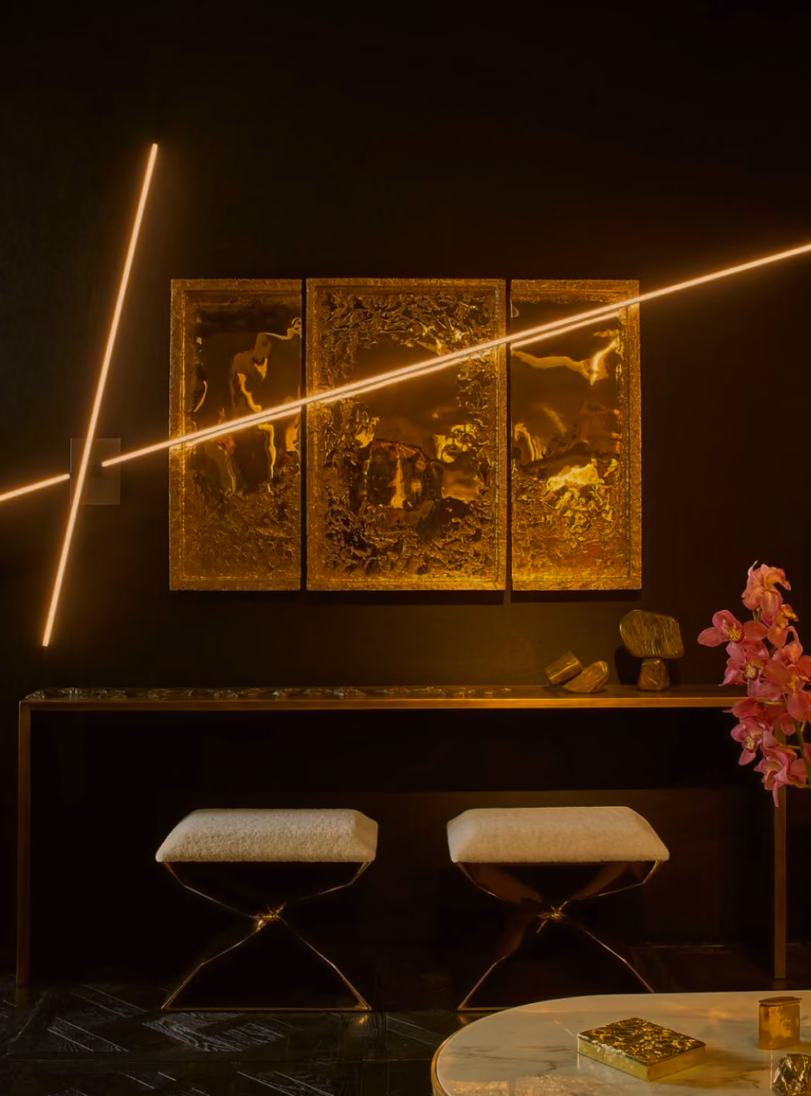 Modern interior with a dark wall featuring a triptych of textured golden panels and diagonal golden light fixtures, a narrow console table with decorative items, two white cushioned stools with metal legs, and a round white table with a small golden box and candle on the right side, next to pink orchids.