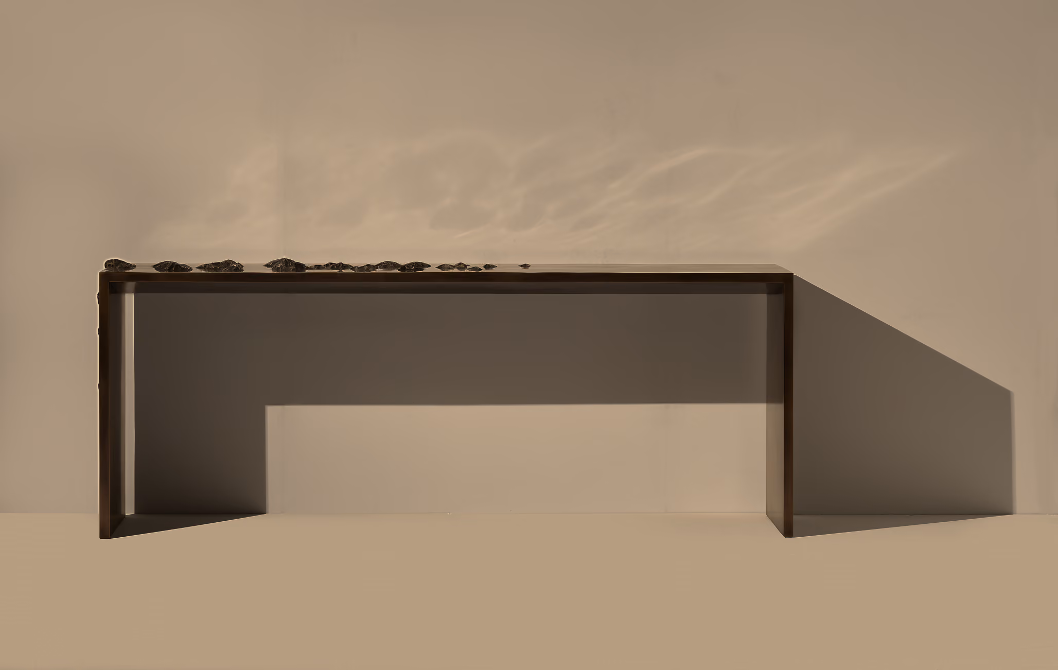 Minimalist dark brown wooden bench casting a long shadow on a beige wall and floor, with small stones arranged in a line on top.
