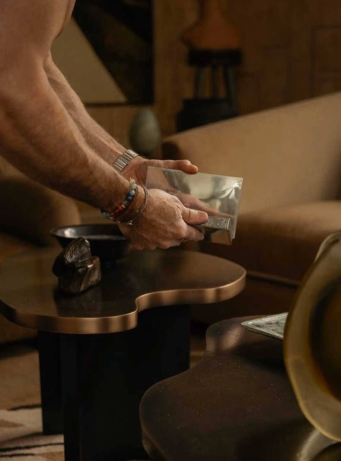 A person wearing bracelets and a watch is holding and examining a metallic rectangular object over a dark wooden coffee table in a cozy living room with a brown couch in the background.