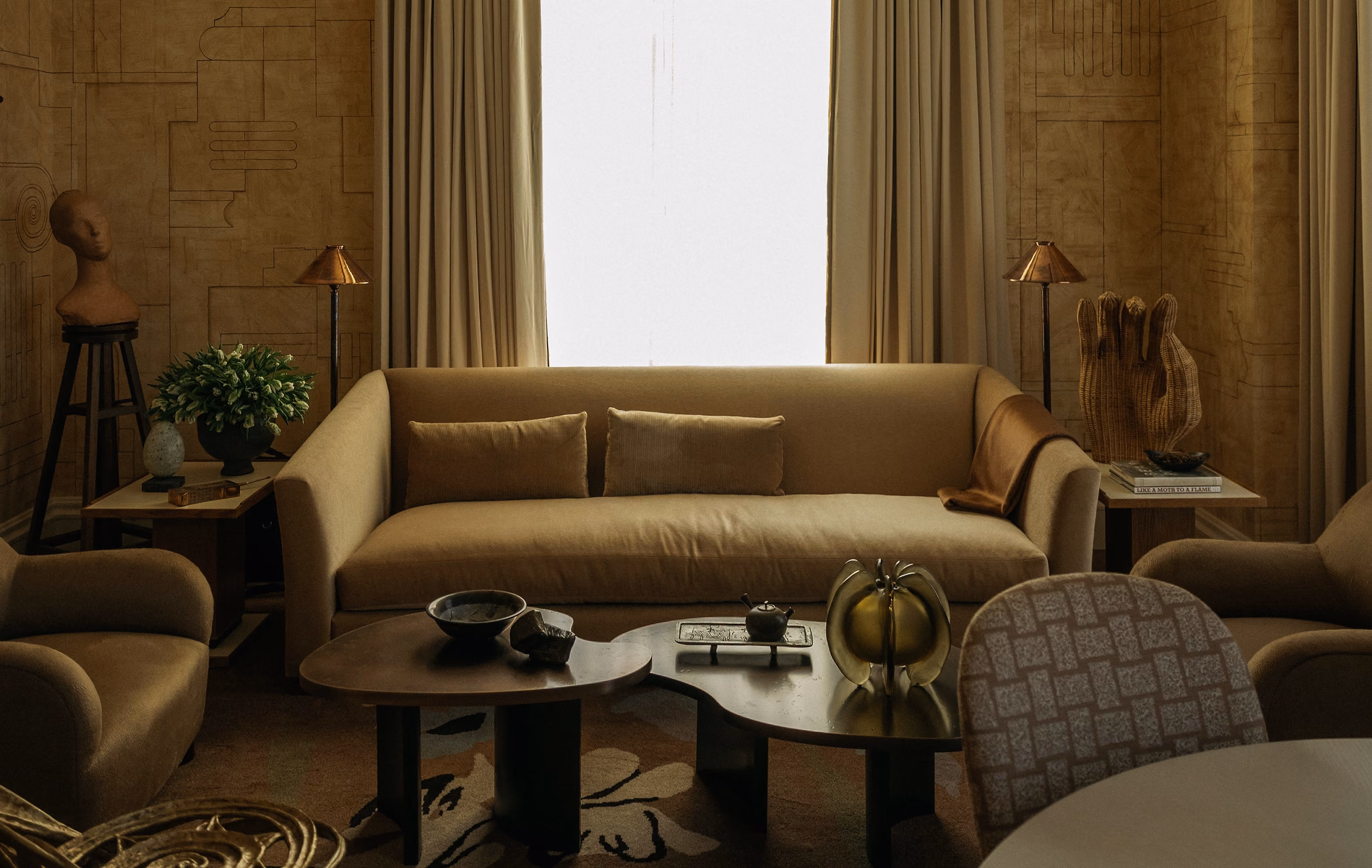 Living room with beige sofa and two matching pillows, flanked by two beige armchairs and side tables with lamps; a medium-size window with curtains behind sofa, abstract wall art, two coffee tables with decorative items, and a patterned chair in foreground.