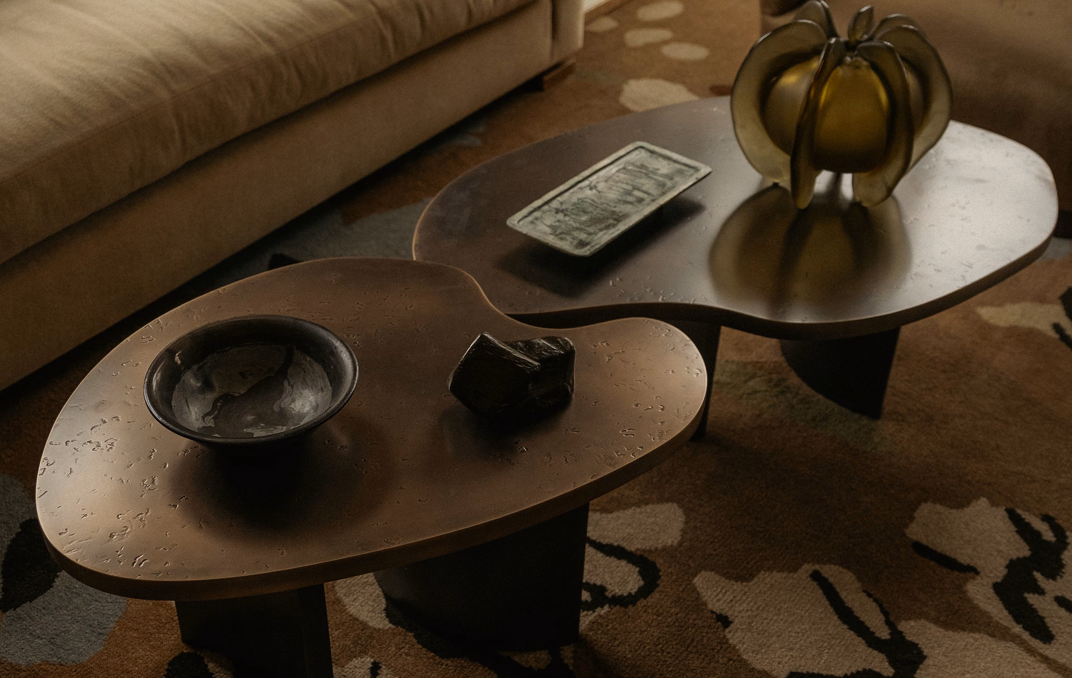 Two dark brown wooden coffee tables with organic, rounded shapes placed on a patterned rug, with a beige couch partially visible in the background; one table holds a bowl and a small dark object, the other has a rectangular dish and a yellow decorative pumpkin.