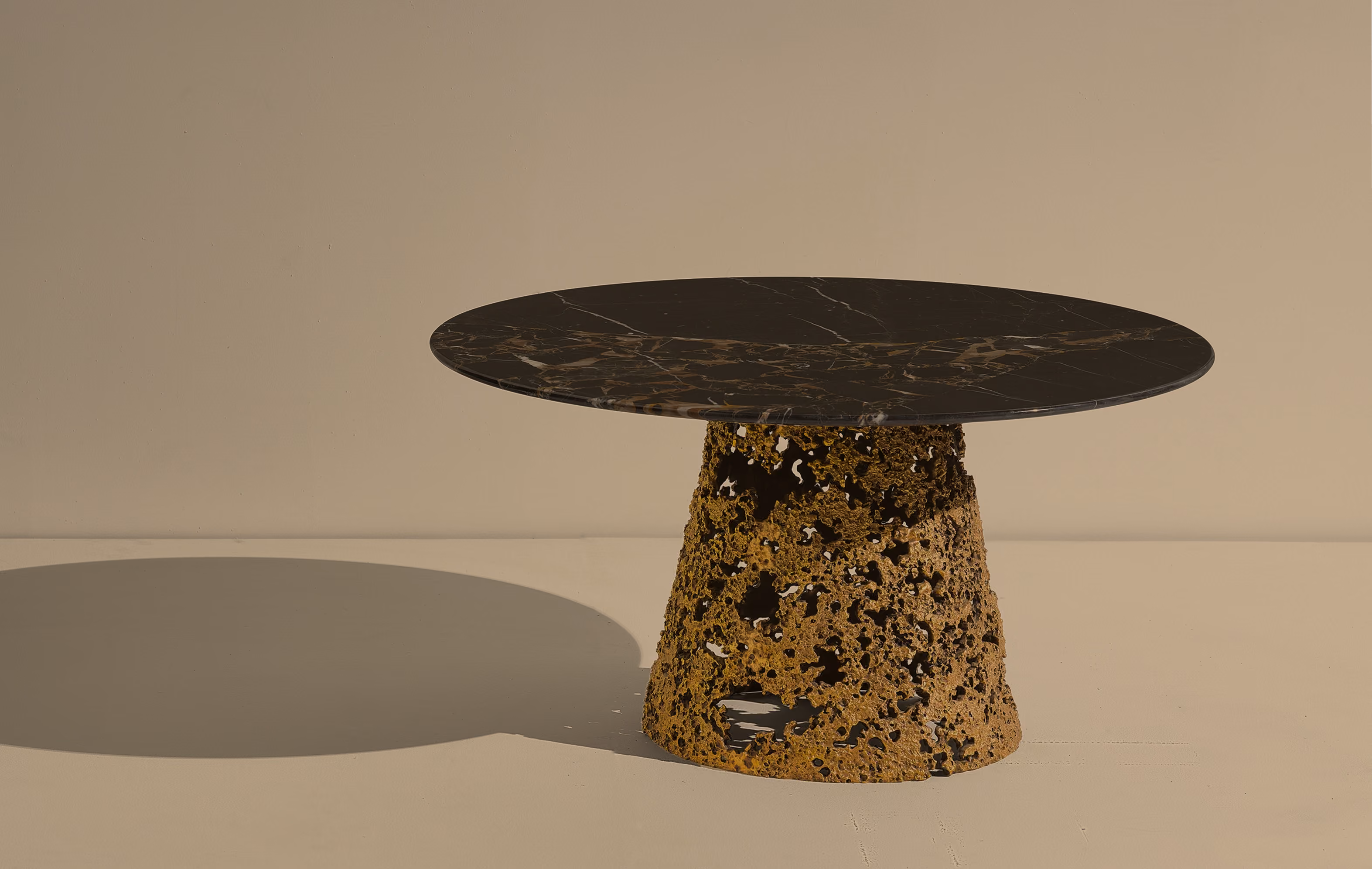 Modern round table with a black marble top and a unique textured golden base casting a shadow on a light beige surface and background.