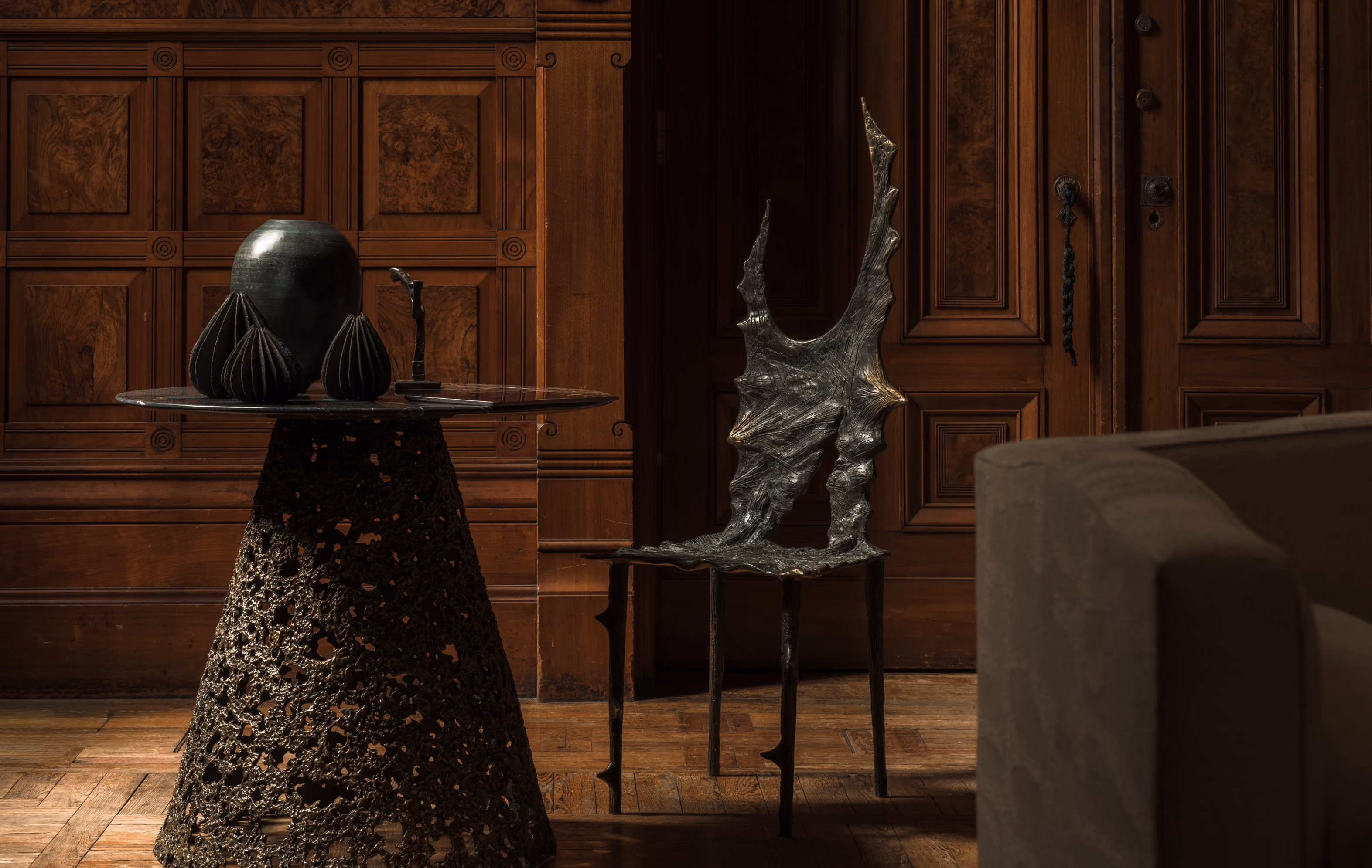 Dark textured metal table and chair with intricate, organic perforated designs set against wooden paneled walls and parquet floor in a dimly lit room.