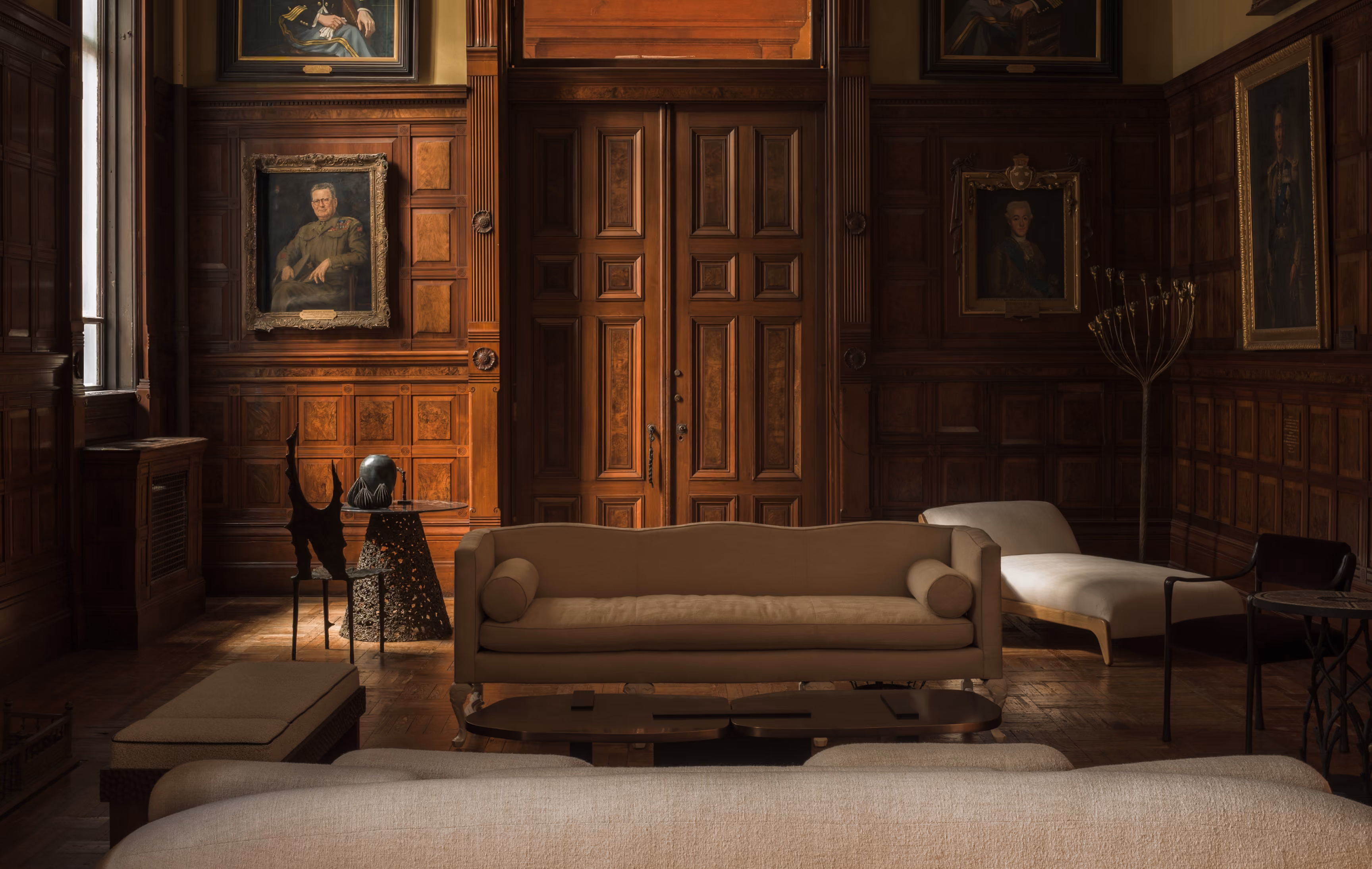 Dimly lit elegant room with wooden paneled walls and large wooden double doors at the center, featuring classic portraits and upholstered beige sofas and chairs arranged around a low coffee table.