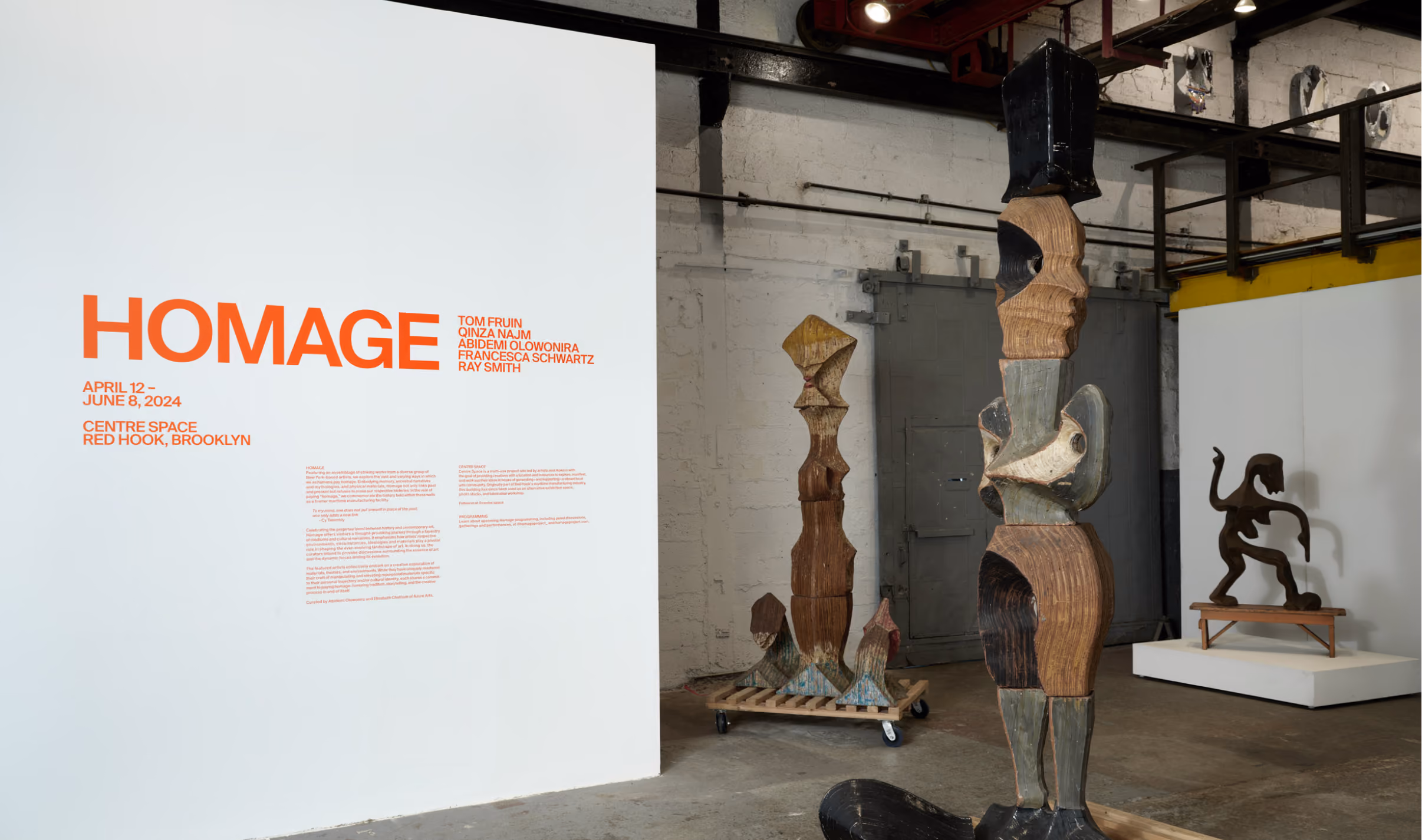 Interior view of an art gallery with a white wall displaying orange text reading HOMAGE, exhibition dates April 12 to June 8, 2024, and location Centre Space, Red Hook, Brooklyn, alongside a list of artist names. In the gallery space are three abstract wooden sculptures with geometric and organic shapes; one tall sculpture with multiple segments is in the foreground, another is on a wheeled platform in the middle, and a smaller sculpture on a stand is in the background.