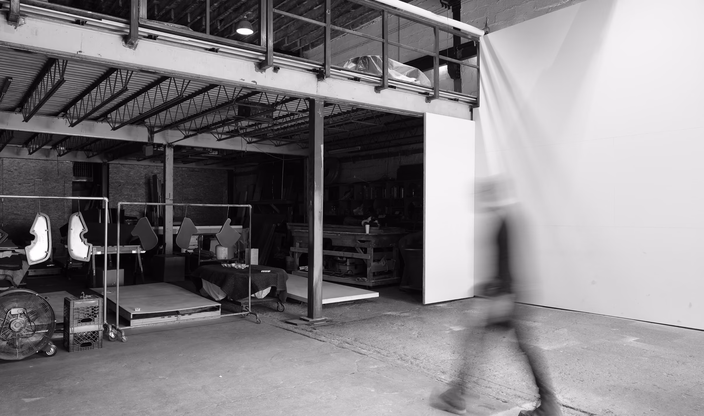 Black and white photo of the interior of a warehouse or workshop with equipment and materials stored on the left side and a blurred figure walking on the right side.