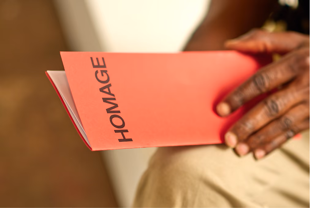 Close-up of a person's hand holding a red booklet with the word 'HOMAGE' printed vertically in black on the cover.