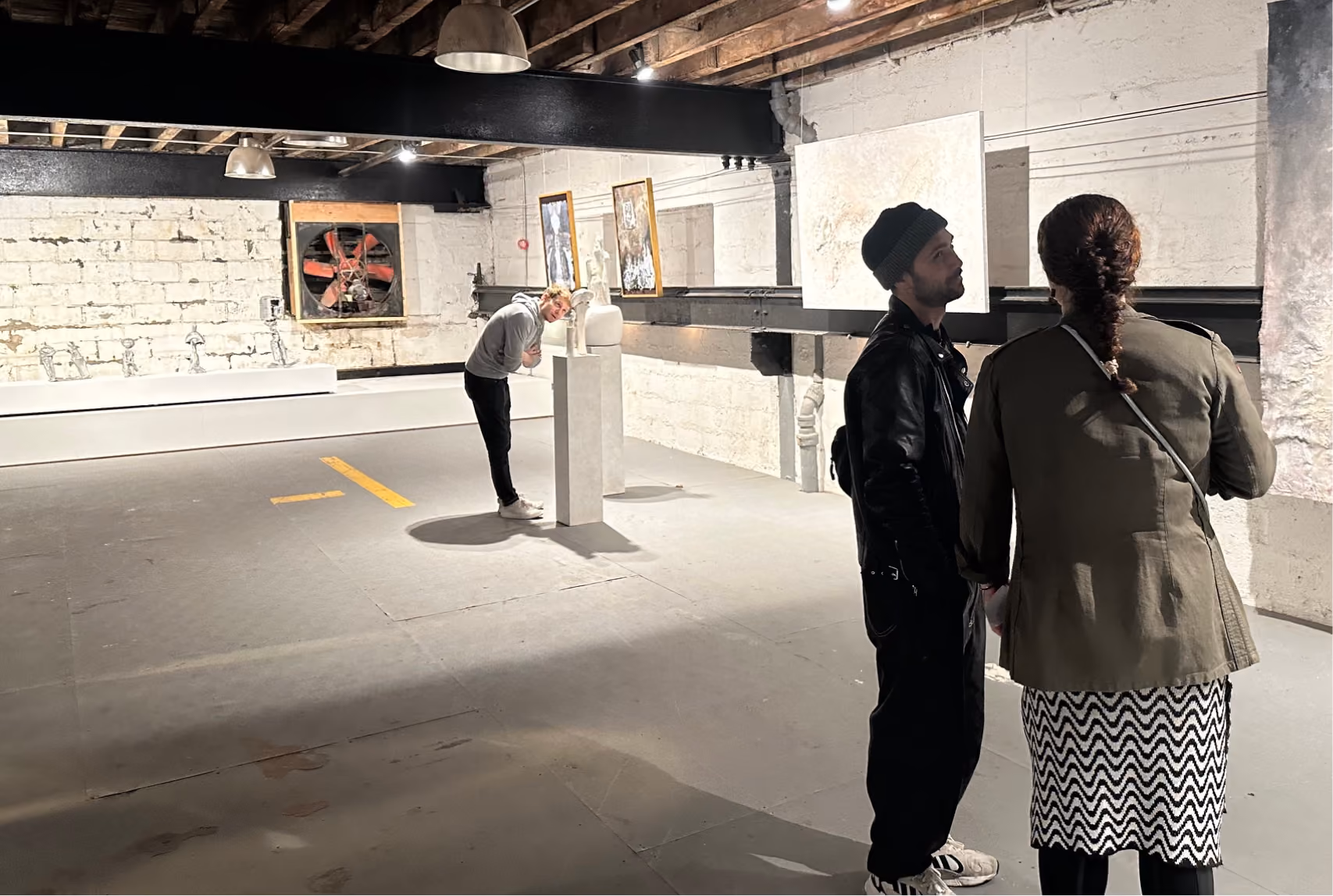 Two people closely examining a large painting on a white brick wall in a dimly lit gallery with exposed wooden ceiling beams, while another person leans in to look at a white sculpture on a pedestal under warm spotlights.