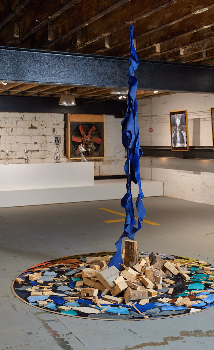 Art installation in an industrial-style gallery with a rough wooden ceiling and concrete floor, featuring a circular arrangement of scattered wooden blocks and colorful fragments on the floor, with a blue twisted fabric hanging vertically from a black metal beam above the center.